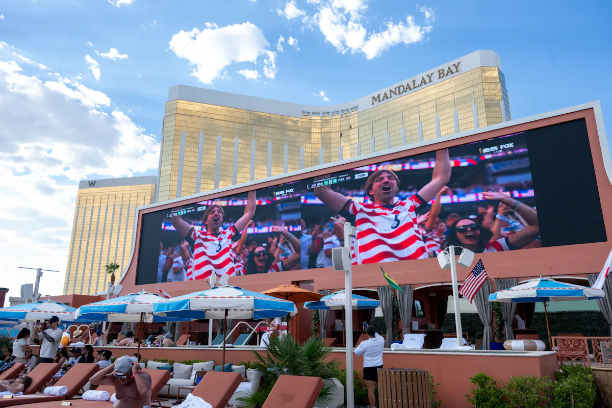 Big screen showing USA fans over packed daybeds, Mandalay Bay behind at Tailgate Beach Club