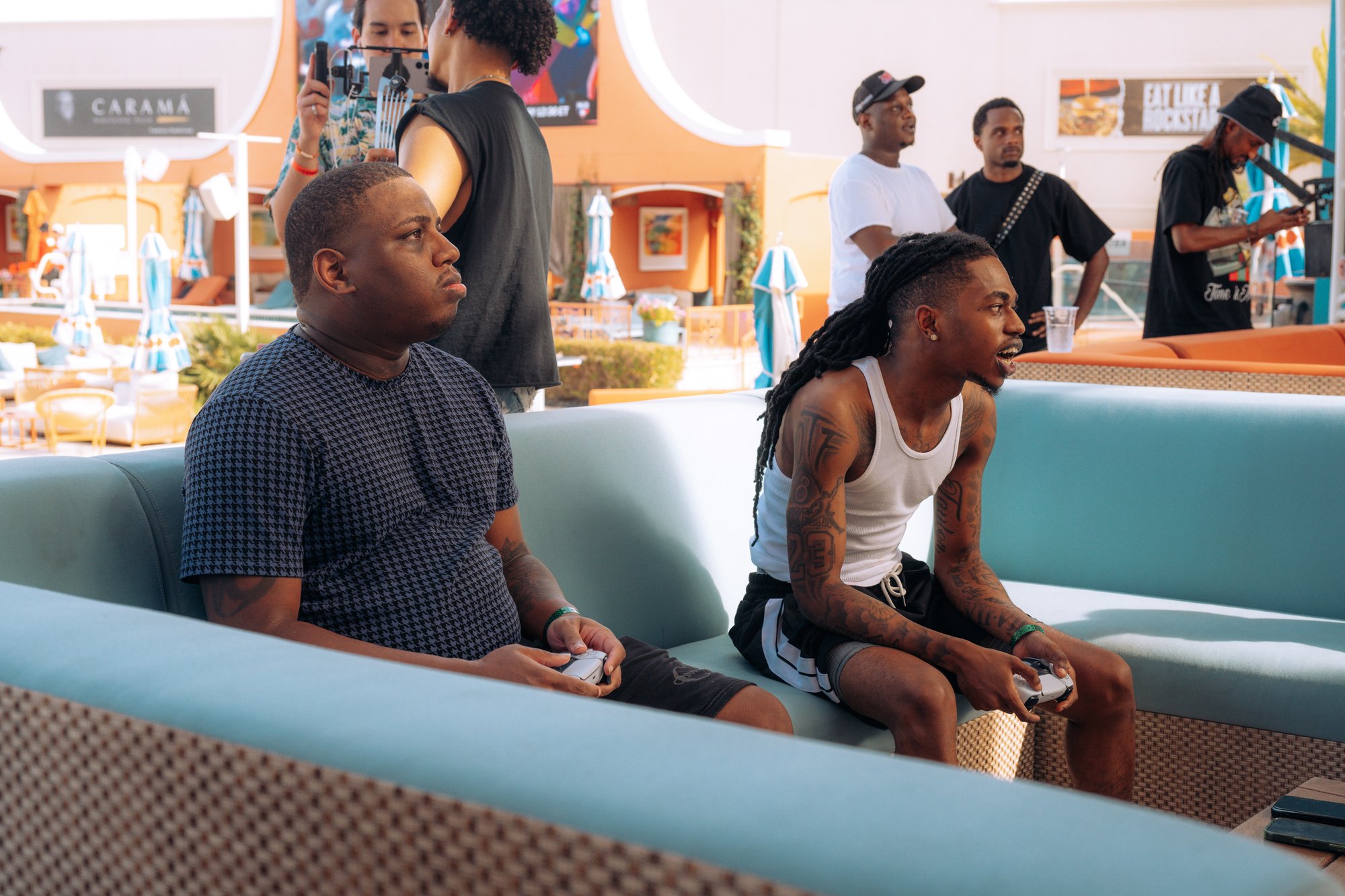 Two players side by side on an aqua daybed at Tailgate Beach Club, controllers in hand and eyes on the game, with the crowd and pool deck behind them