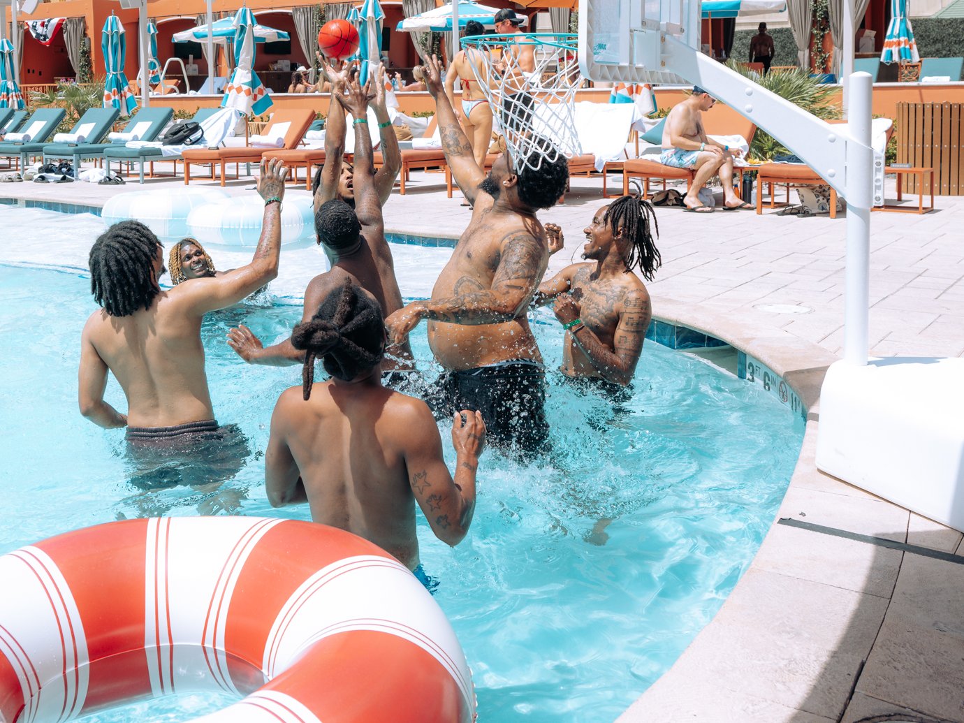Guests in the pool at Tailgate Beach Club jump for a ball under the pool hoop, a red-and-white striped float in front and lounge chairs behind