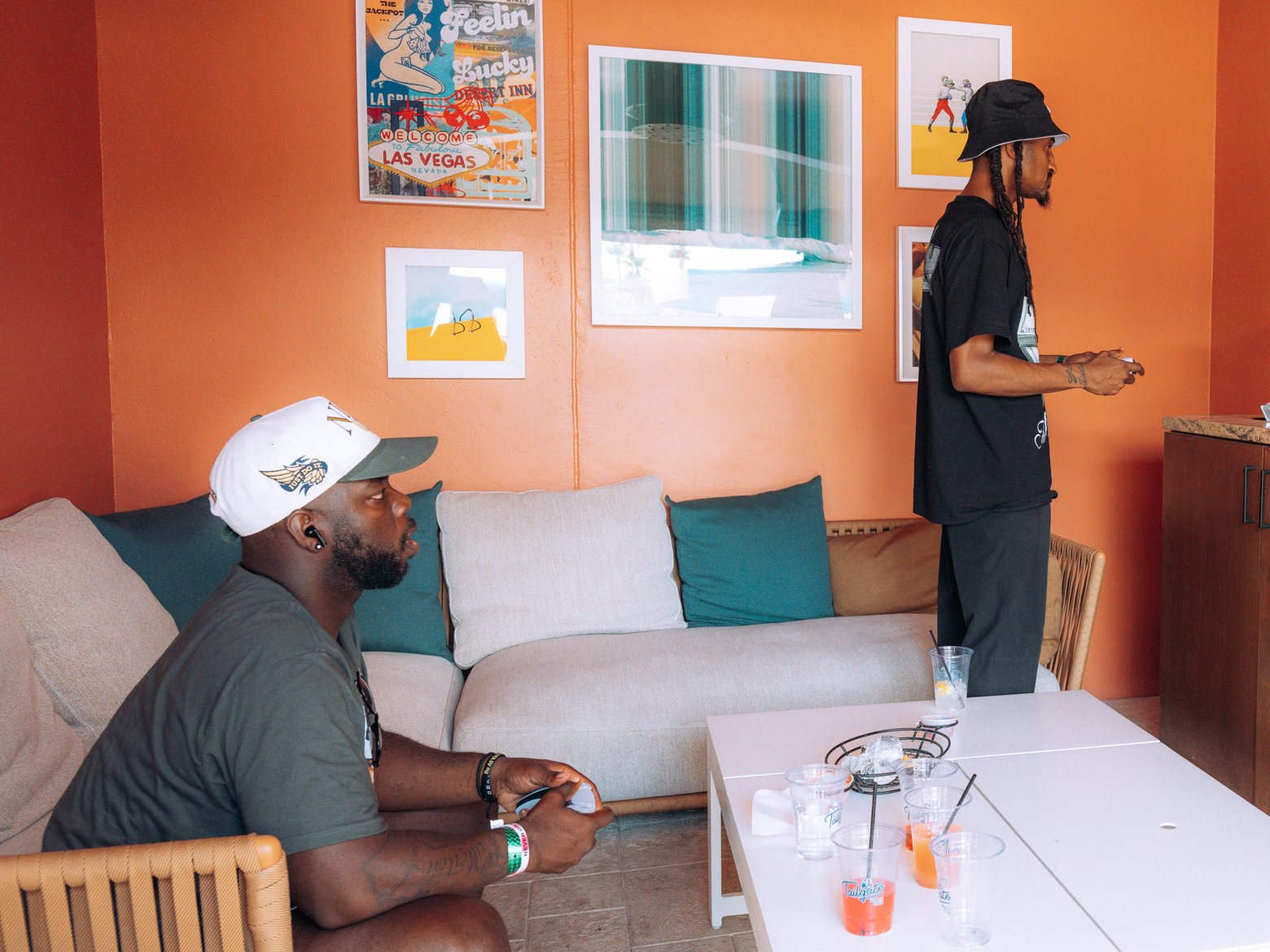 Inside an orange Tailgate Beach Club cabana, a player in a white cap sits on the couch with a game controller while another player stands by the counter
