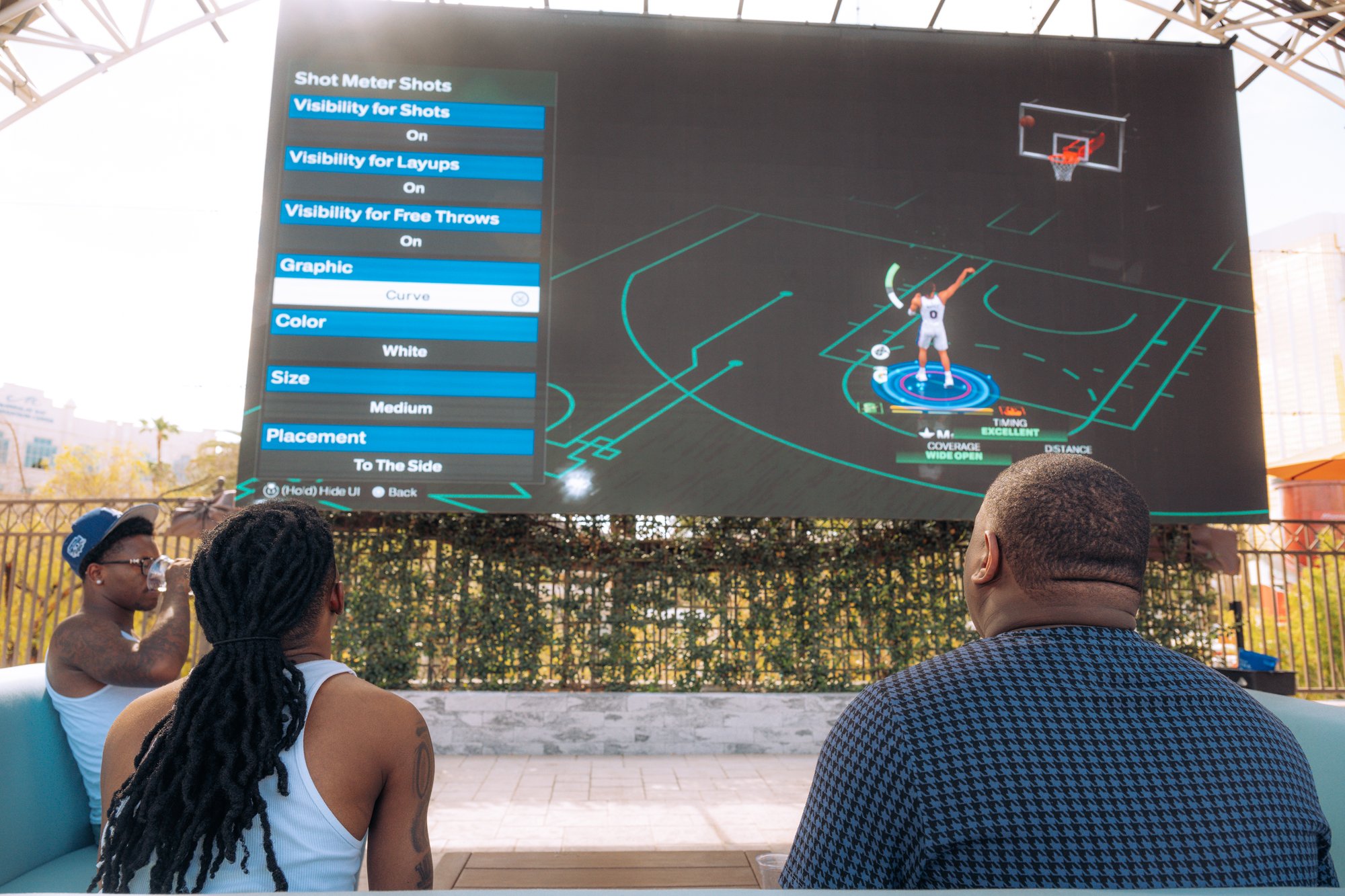 Two players seated on a poolside daybed at Tailgate Beach Club look up at a giant LED screen showing video game settings under the covered stage canopy