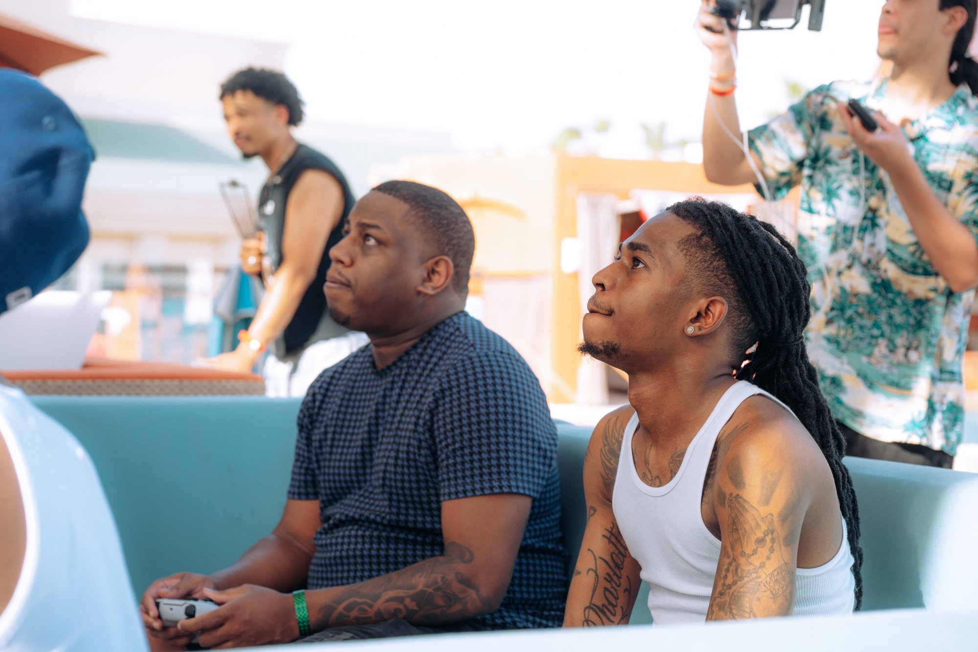 Two players on a daybed with controllers watch the screen while guests mingle behind them on the pool deck