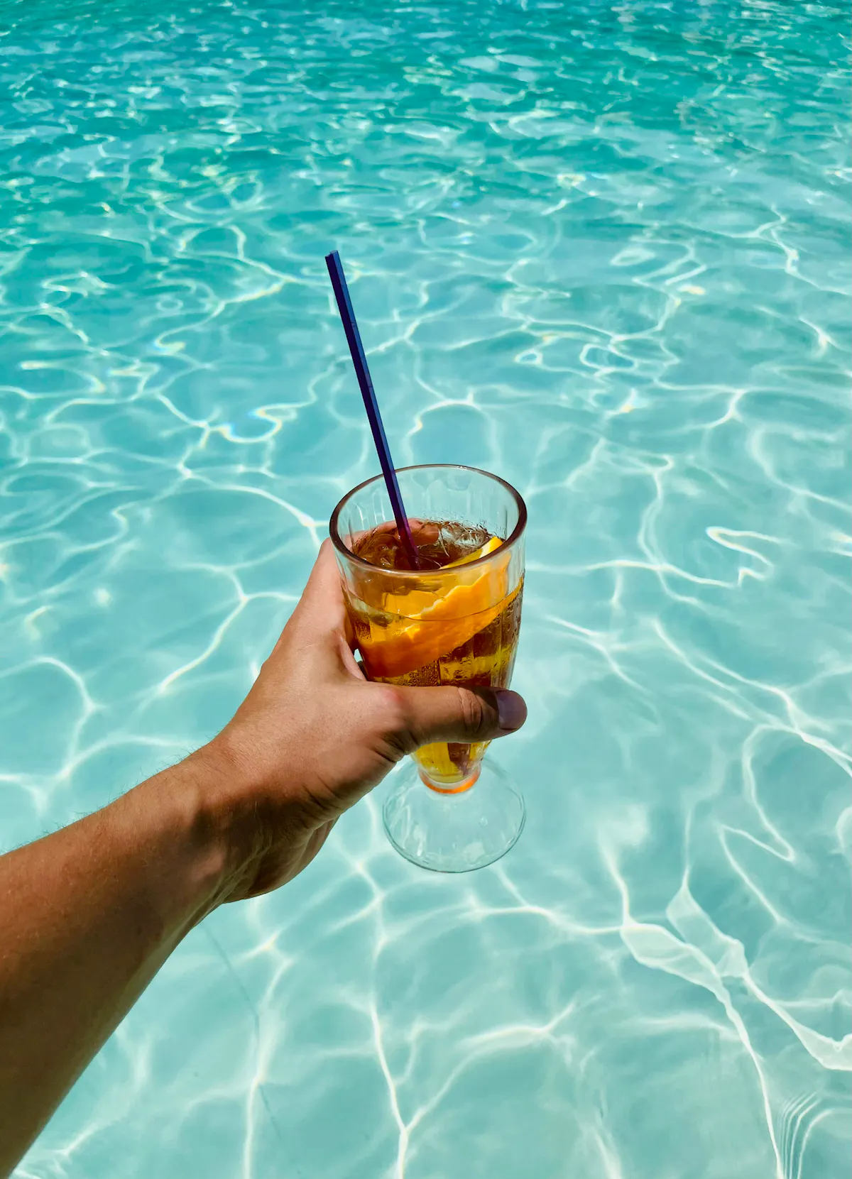 Hand holding cocktail over turquoise pool water at Tailgate Beach Club