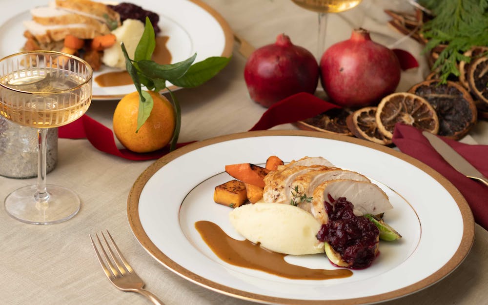 Carved turkey on a gold trimmed plate with mashed potatoes, cranberry sauce, gravy, carrots, and a glass of champagne to the left. On the right of the plate can you see festive decor such as dried oranges, pomergrantes.