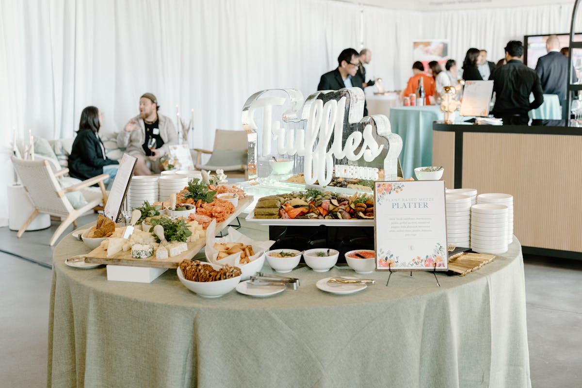 Elegant catering display at Truffles Fine Foods’ Spring Fling 2025, featuring plant-based mezze, cheese and charcuterie boards, dips, and crackers arranged around an ice sculpture with the Truffles logo, set in a bright lounge-style venue at VanDusen Botanical Garden