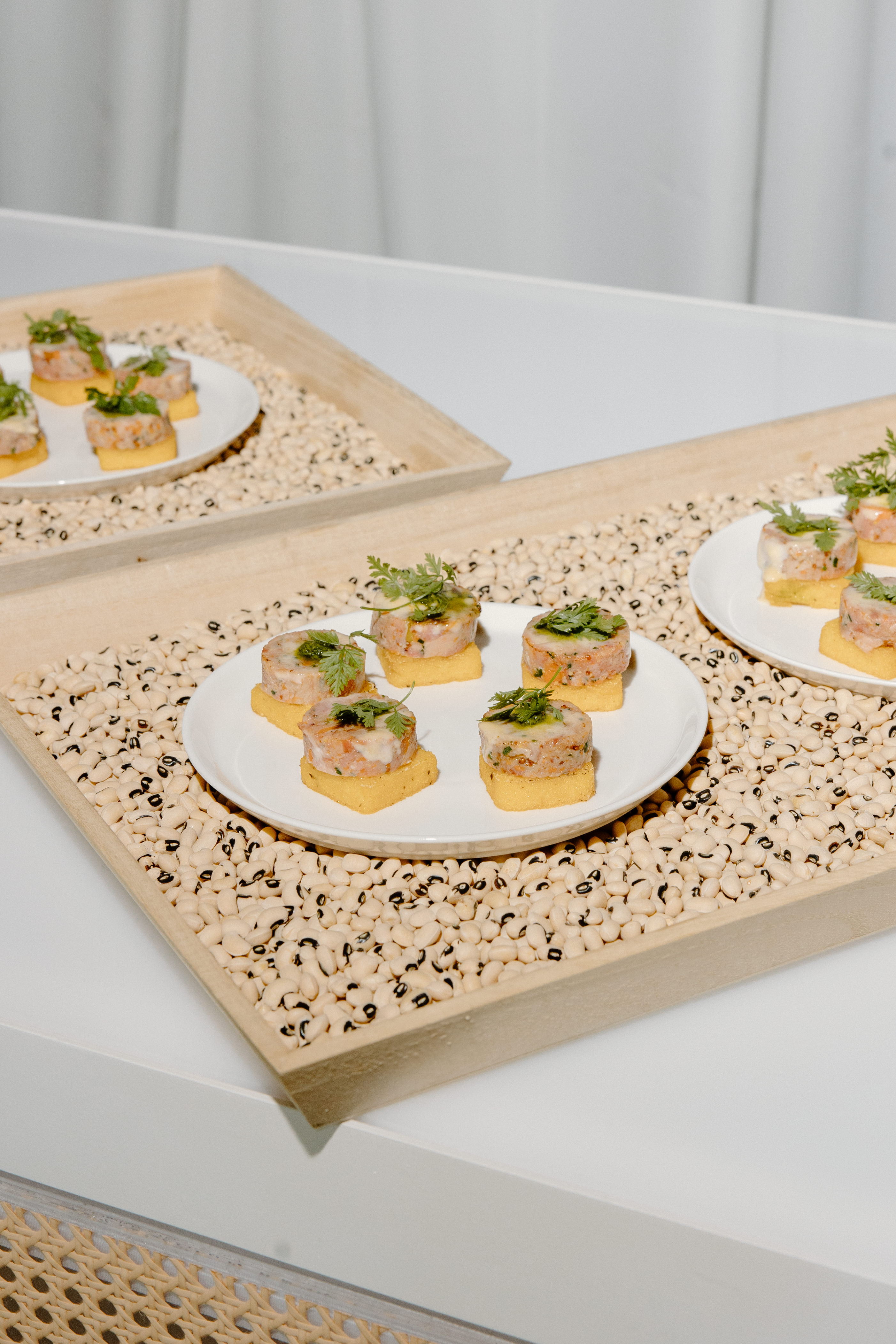 Smoked pork hock appetizer bites served on polenta squares and garnished with fresh herbs, elegantly plated on white dishes over trays filled with black-eyed peas at Truffles Fine Foods' Spring Fling 2025 event in Vancouver.