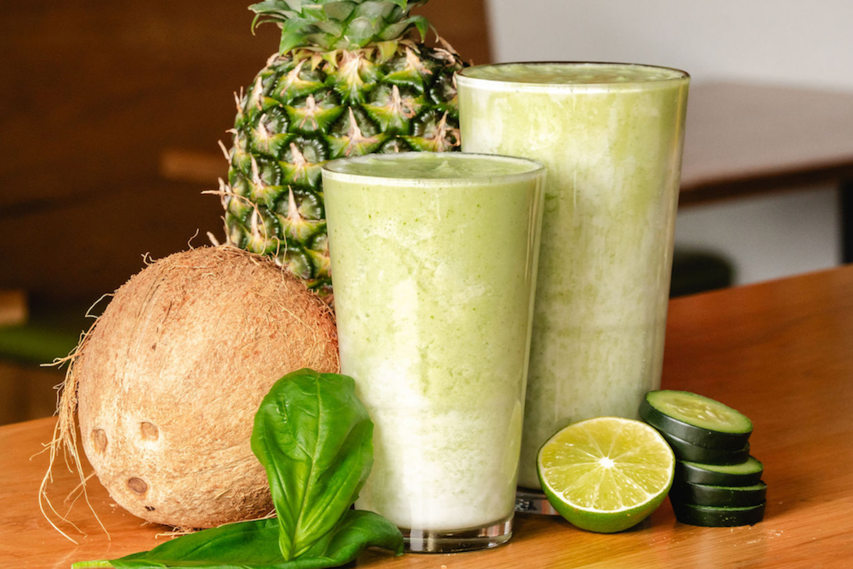 a glass of green smoothie next to a pineapple and coconut