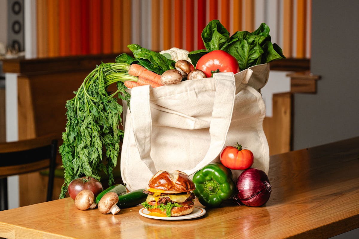 Sunrise Burger sits on a wooden table in front of a reusable canvas grocery bag filled with fresh organic vegetables, including carrots, bell peppers, tomatoes, mushrooms, and onions. The burger is stacked with a Beyond™ patty, vegan Yo-Egg, tempeh bacon, lettuce, tomato, and melted cheese on a toasted pretzel bun. The scene highlights fresh, organic ingredients and sustainable eating.