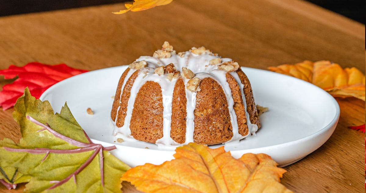 Pumpkin spice mini bundt cake drizzled with white icing and topped with chopped walnuts, served on a white plate. The cake is surrounded by colorful autumn leaves on a wooden table, creating a festive fall atmosphere.