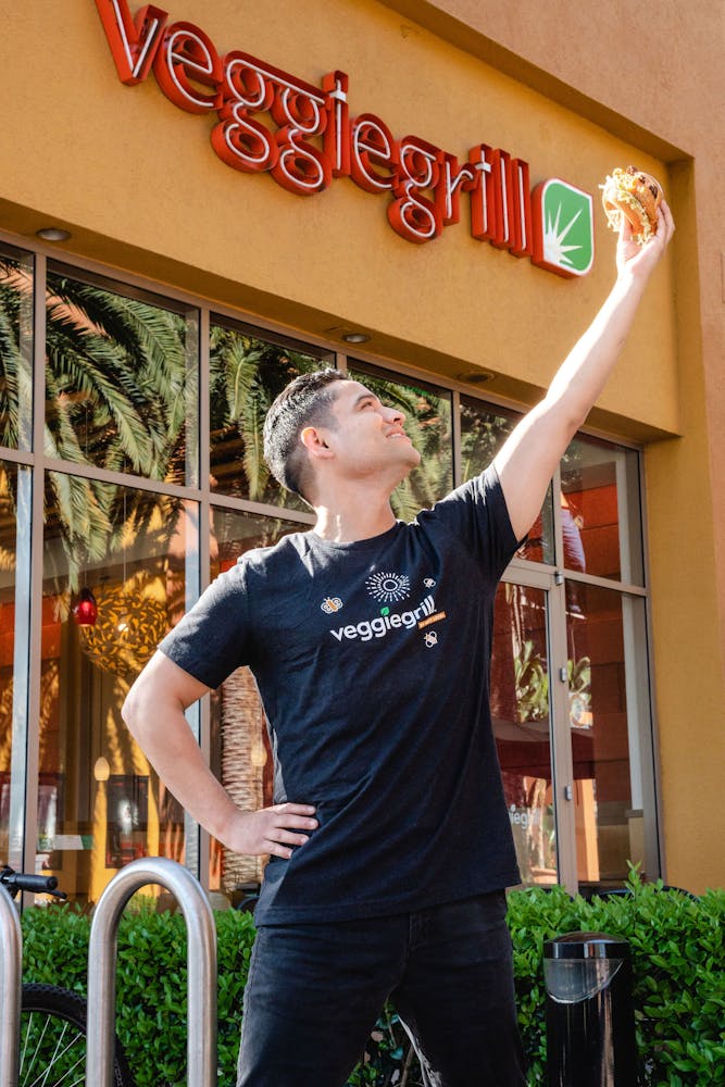 a person holding a burger into the air next to a Veggie Grill store like a superhero