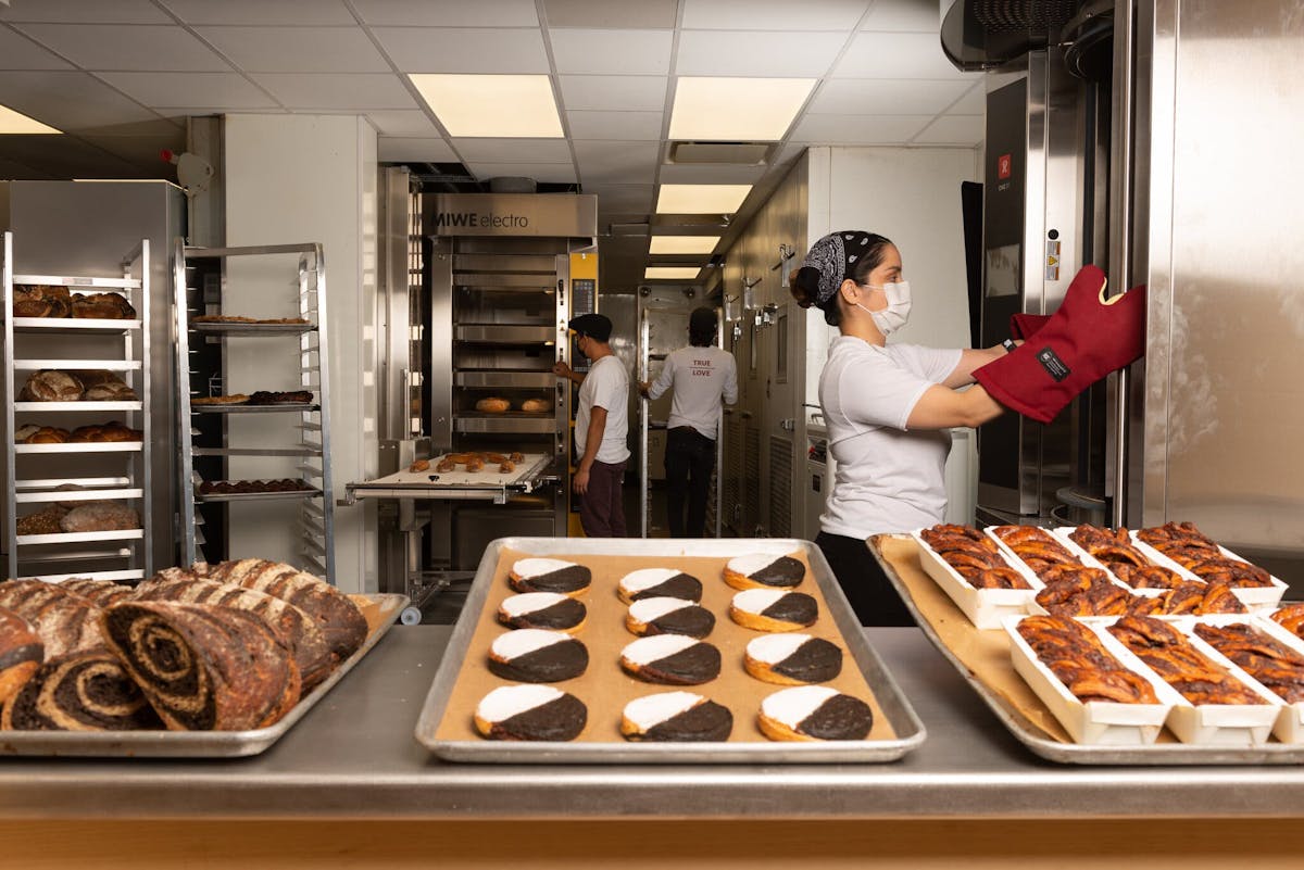 A tray of black and white cookies sits on a stainless steel counter in front of a kitchen full of bakers baking