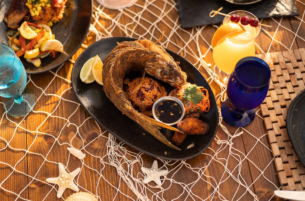 A wooden table decorated with a white fishing net, seashells, and starfish holds a black plate with a whole deep-fried fish served in a curled position. The dish is accompanied by a mound of seasoned rice and beans, fried plantains, shredded carrot garnish, lime and lemon wedges, and a small cup of dipping sauce. Surrounding the plate are colorful drinks in blue and clear glasses, along with another plate of grilled chicken and sautéed vegetables in the background.