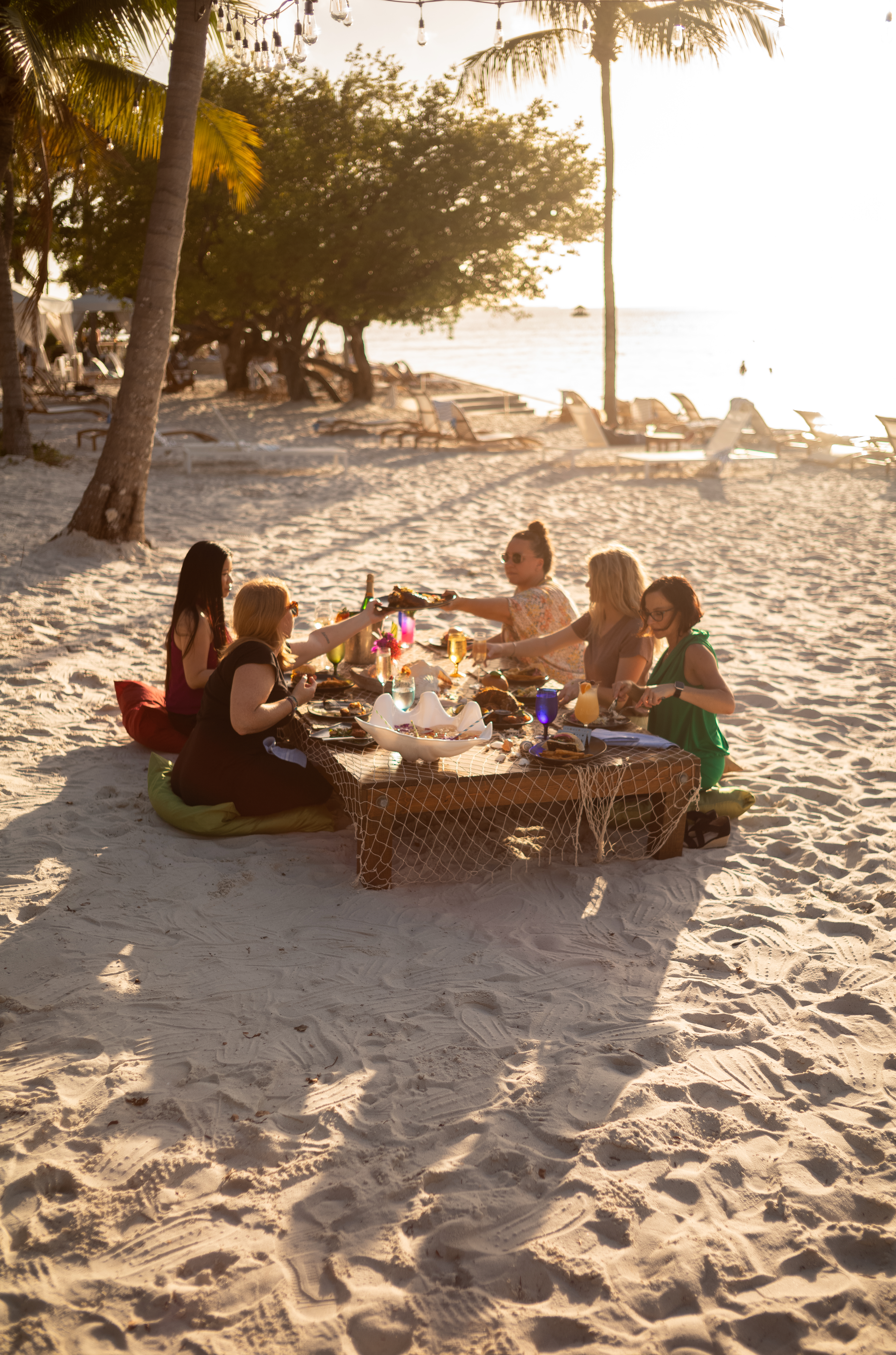 Group of women enjoying a beachfront dinner at a low table in the sand beneath string lights and palm trees.