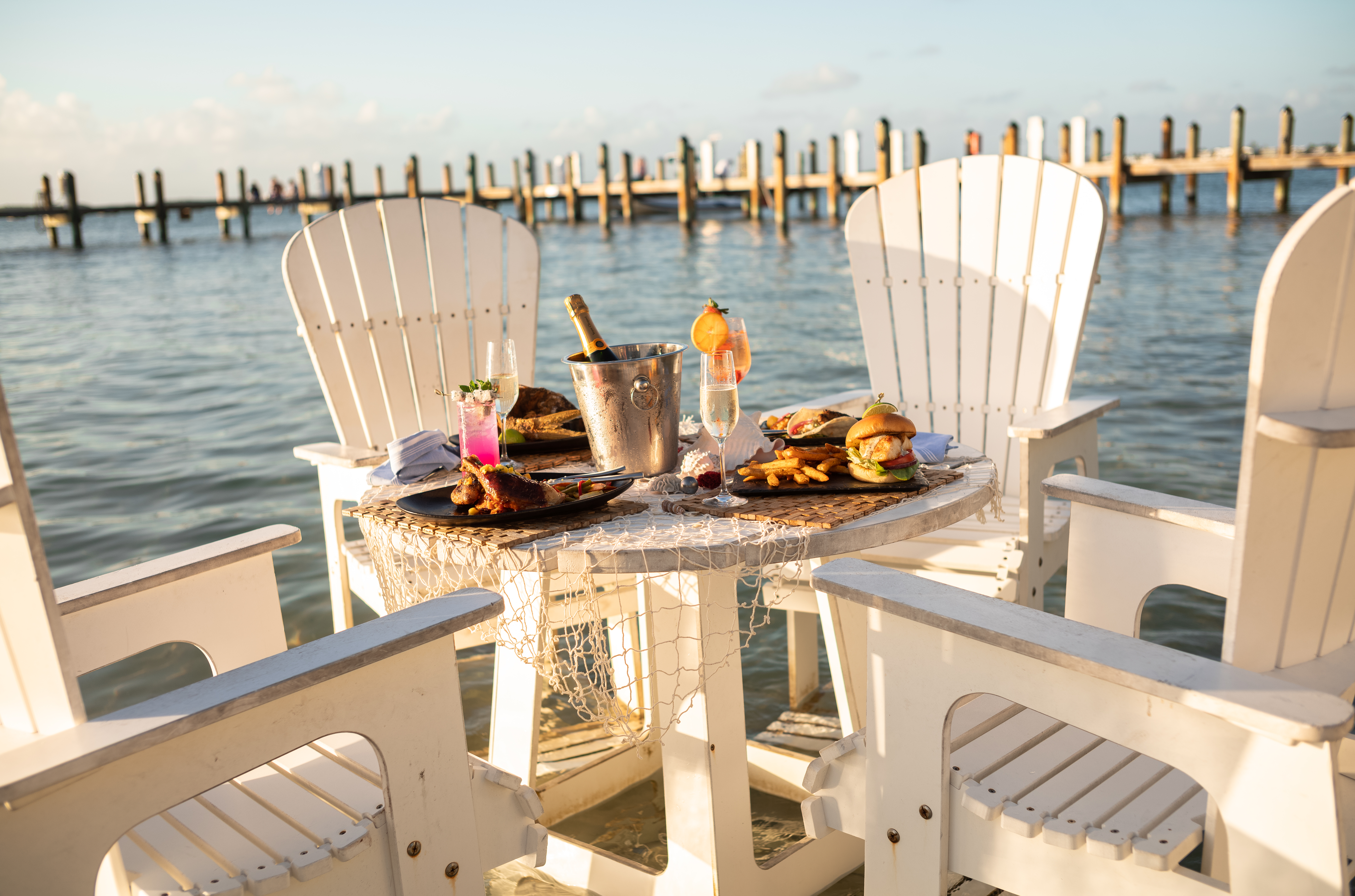 Adirondack chairs surround a table of burgers, fries, and champagne with waves lapping just beneath the dock.