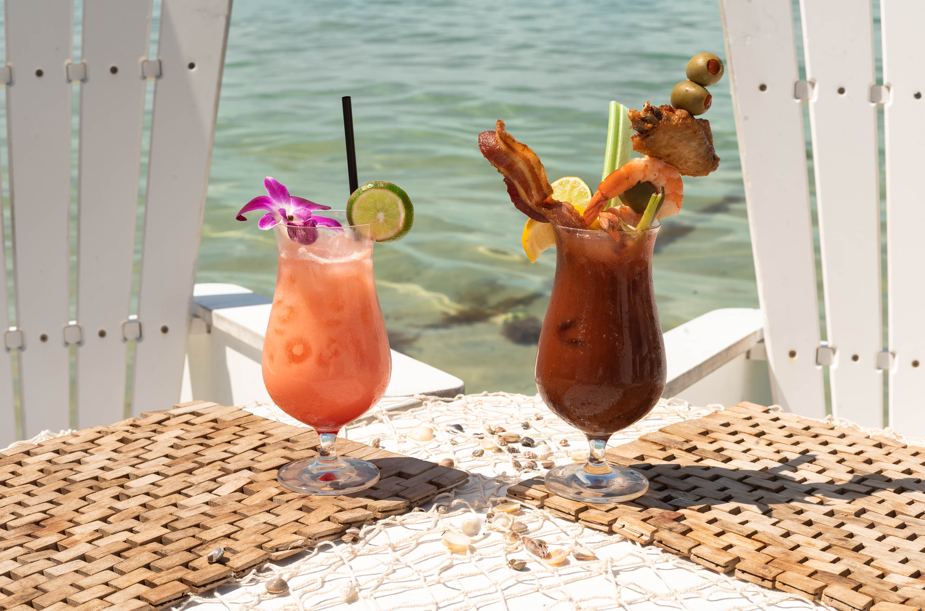 Two tropical cocktails—a pink drink with orchid and lime, and a loaded Bloody Mary—by the water.