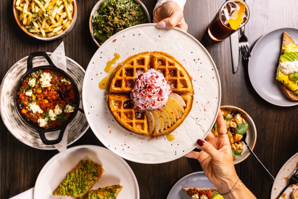 Mochi waffle topped with whipped cream and sliced pear, held above a table of brunch dishes at PLANTA.