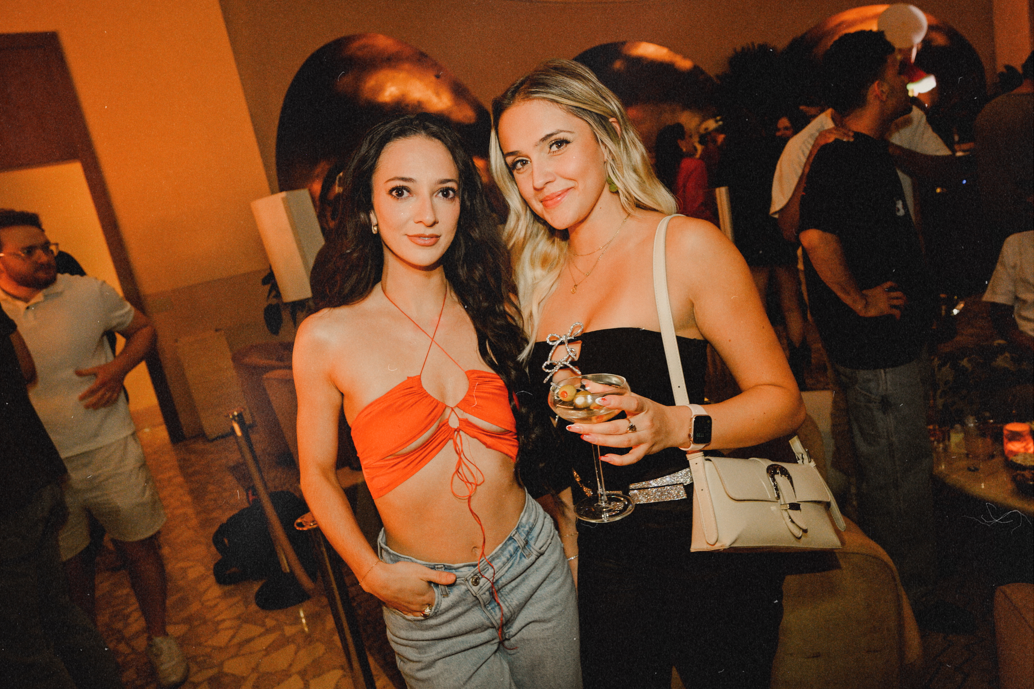 Two women posing together inside a dimly lit lounge at Bel-Aire Lounge, holding cocktails and dressed for a night out with a lively crowd in the background.