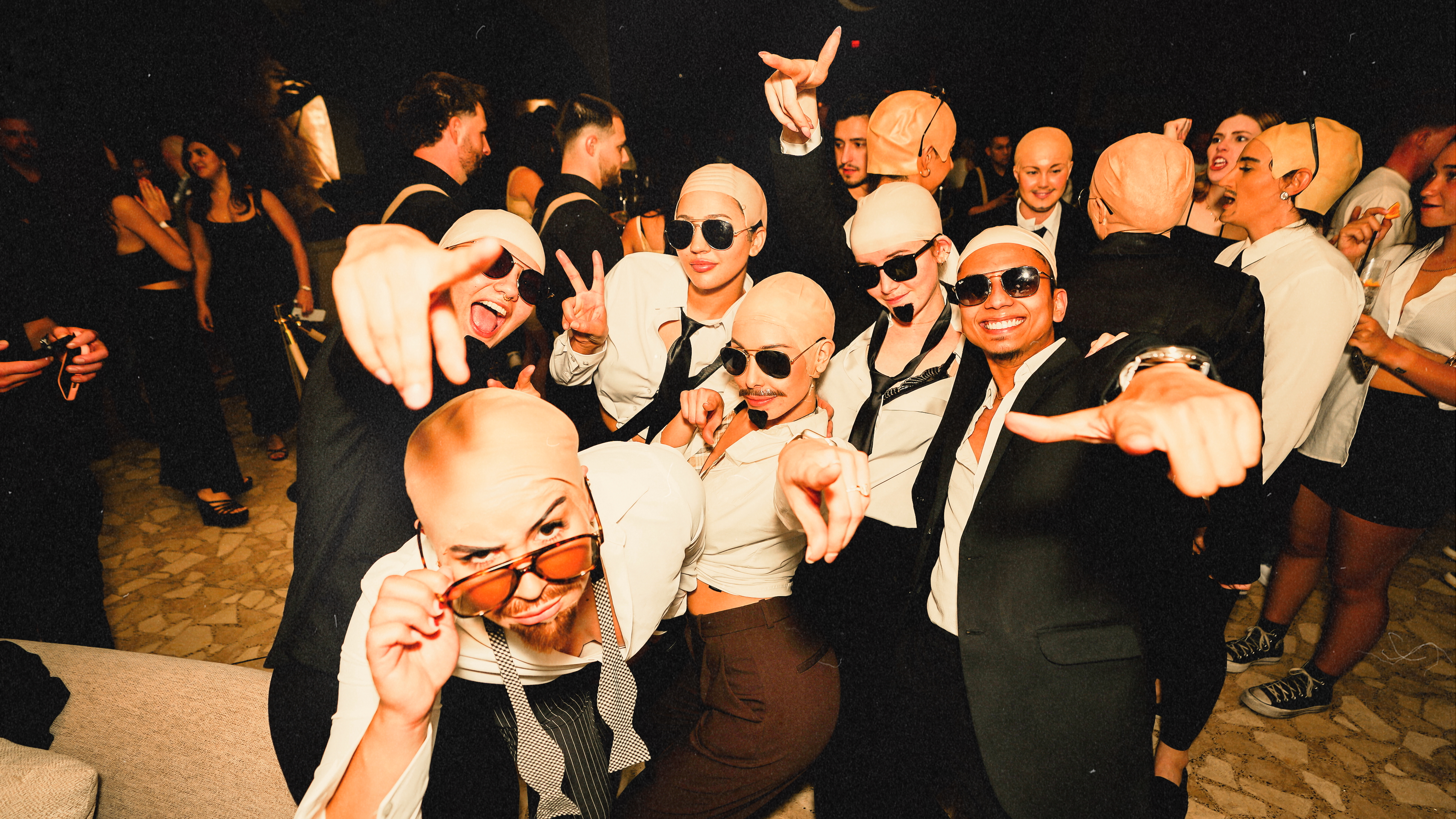Guests dressed in matching Pitbull costumes—bald caps, sunglasses, and ties—celebrate on the dance floor at Bel-Aire Lounge during Baldies Worldwide 2, a packed lounge with DJ event in Las Vegas.