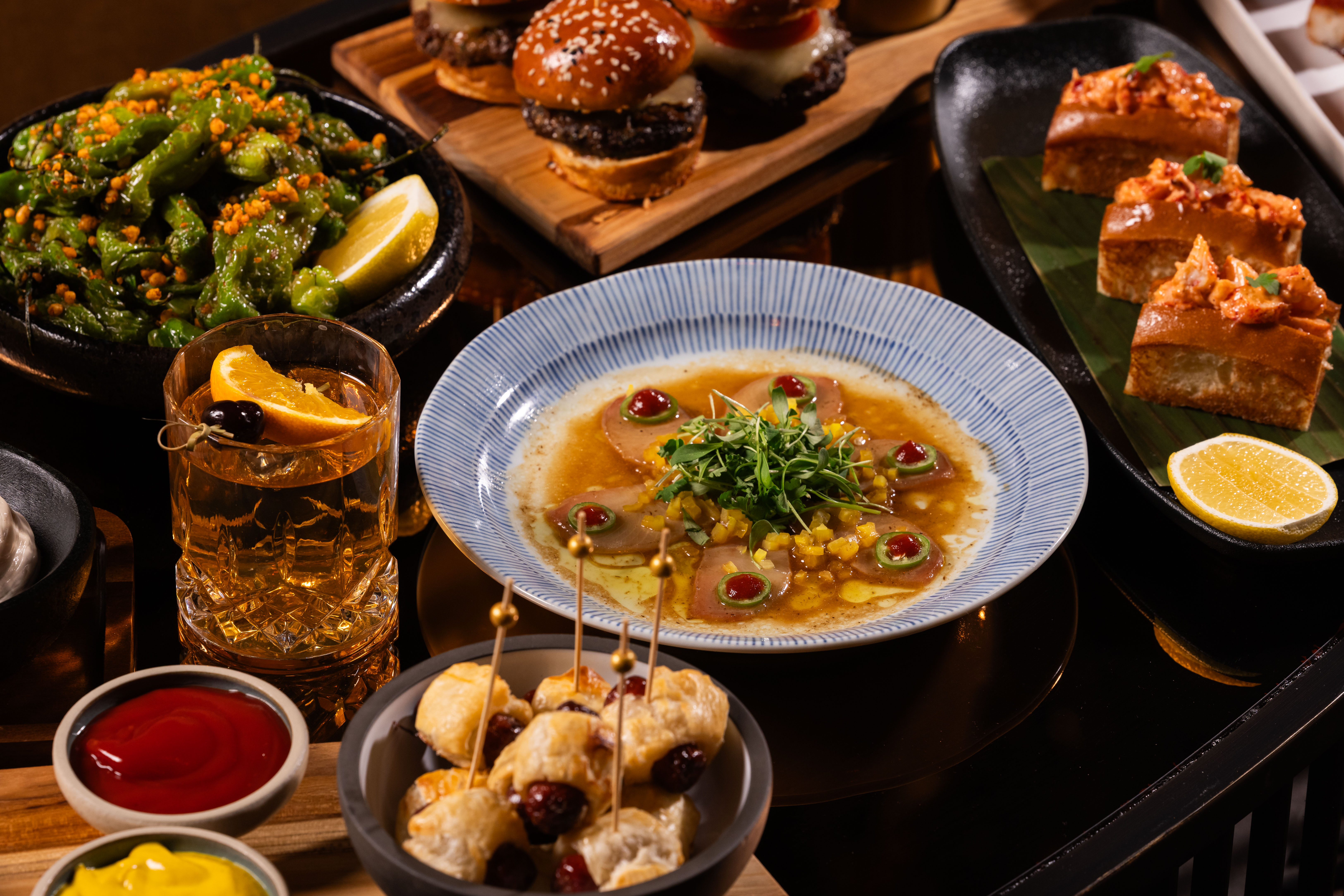 Table snacks at Clique Bar & Lounge in Las Vegas, including wagyu sliders, mini lobster rolls, yellowtail sashimi, and shishito peppers with an Old Fashioned