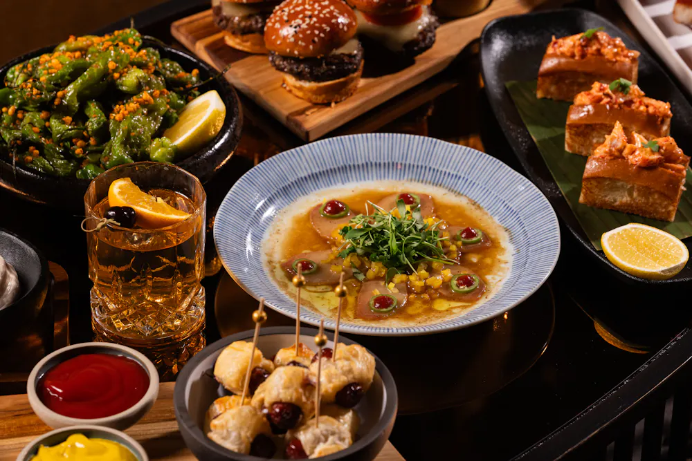 Table snacks at Clique Bar & Lounge in Las Vegas, including wagyu sliders, mini lobster rolls, yellowtail sashimi, and shishito peppers with an Old Fashioned