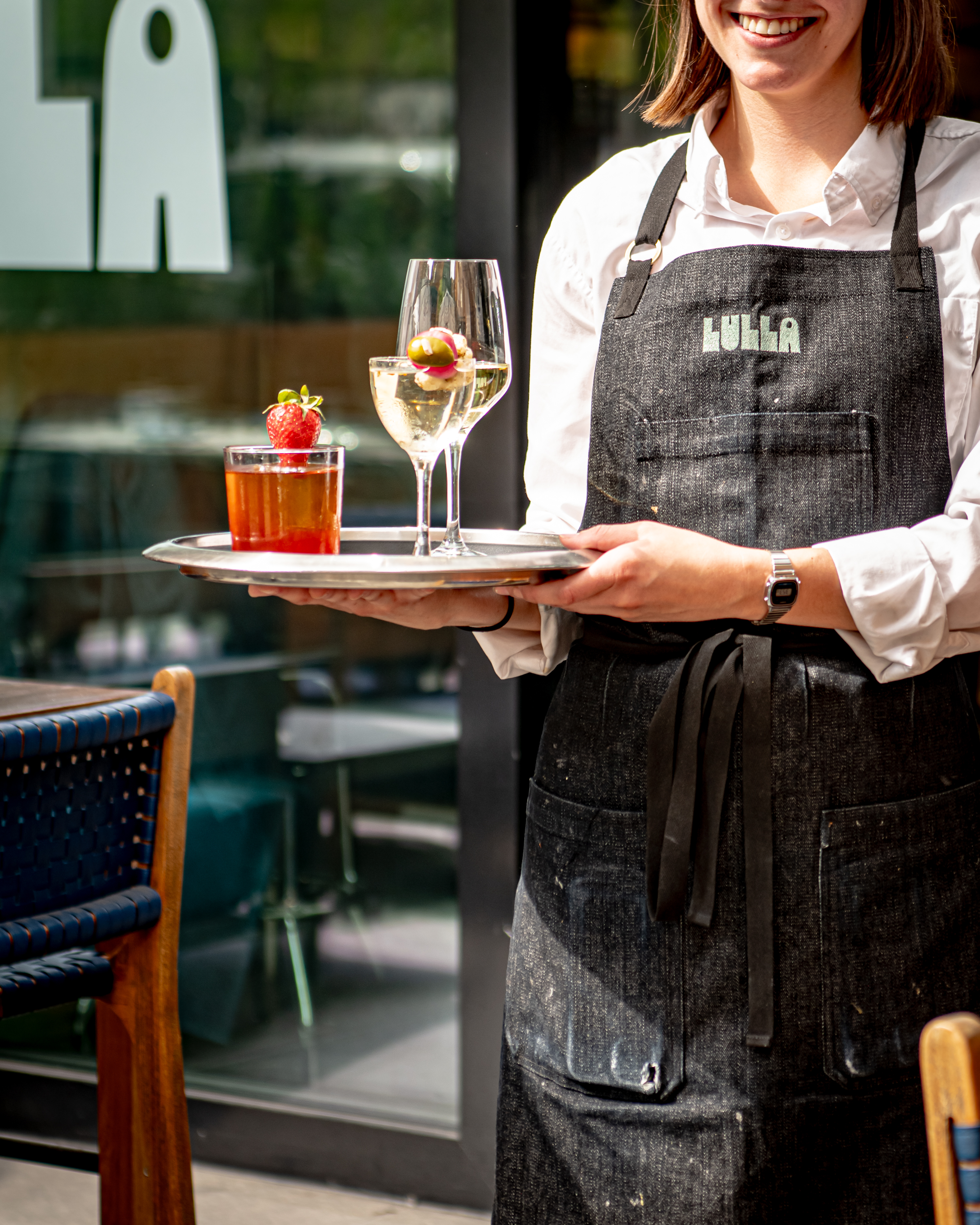 A woman in an apron presents a tray filled with various drinks, showcasing her hospitality and service skills