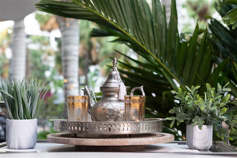 Traditional Moroccan tea service with ornate silver teapot and glassware on a decorative tray, set beside lush palm leaves in a bright dining space.
