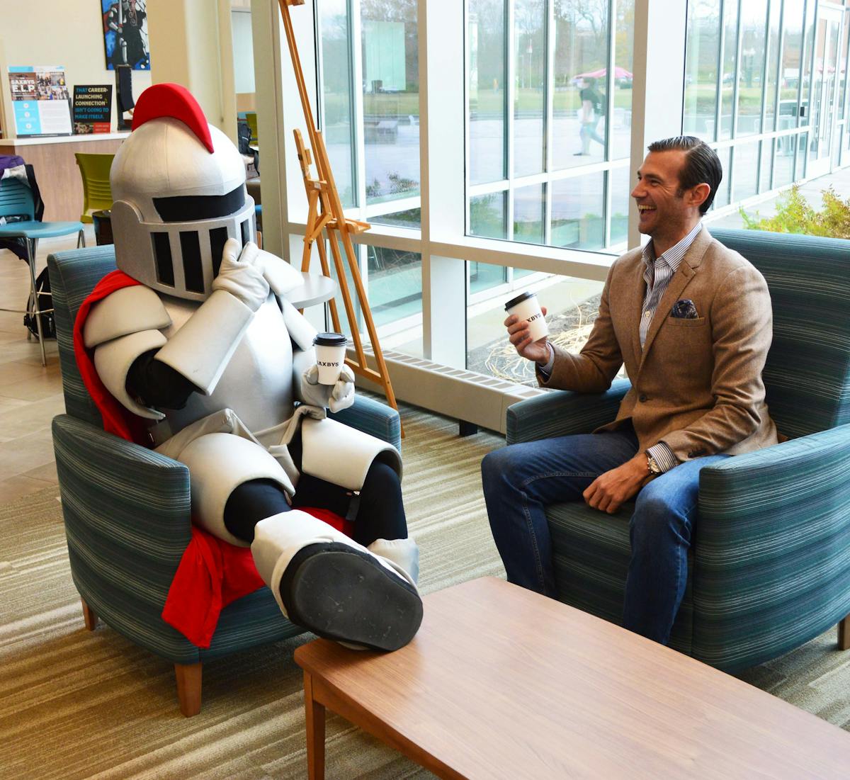 RCBC mascot, Baron, and Saxbys CEO, Nick Bauer sit on two chairs and laugh. Both holding a cup of Saxbys coffee