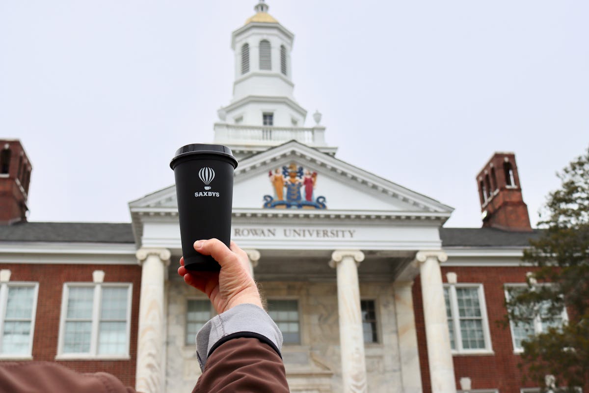 a person holding a Saxbys cup in the air in front of a building