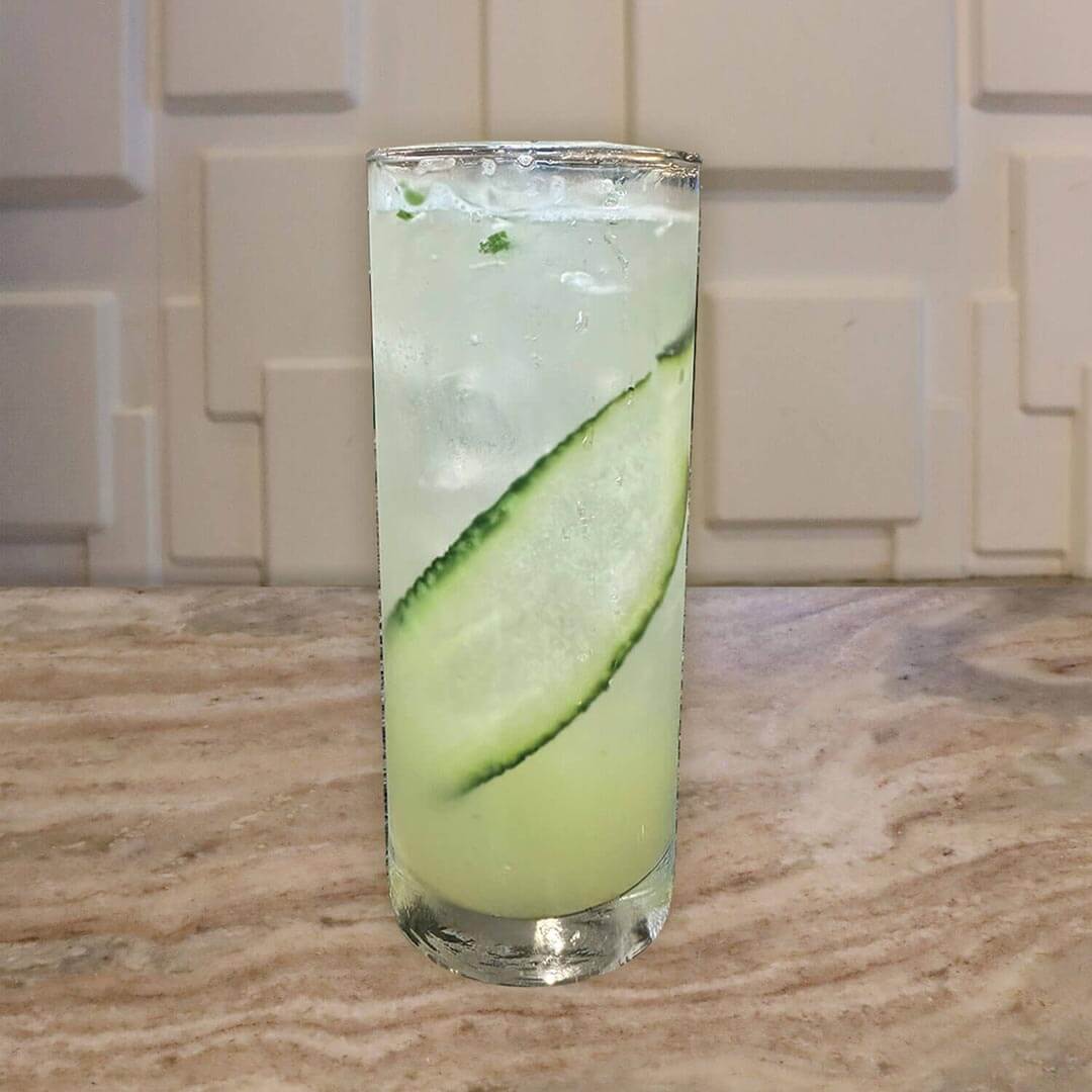 A tall refreshing clear drink with long cucumber slices inside the glass.