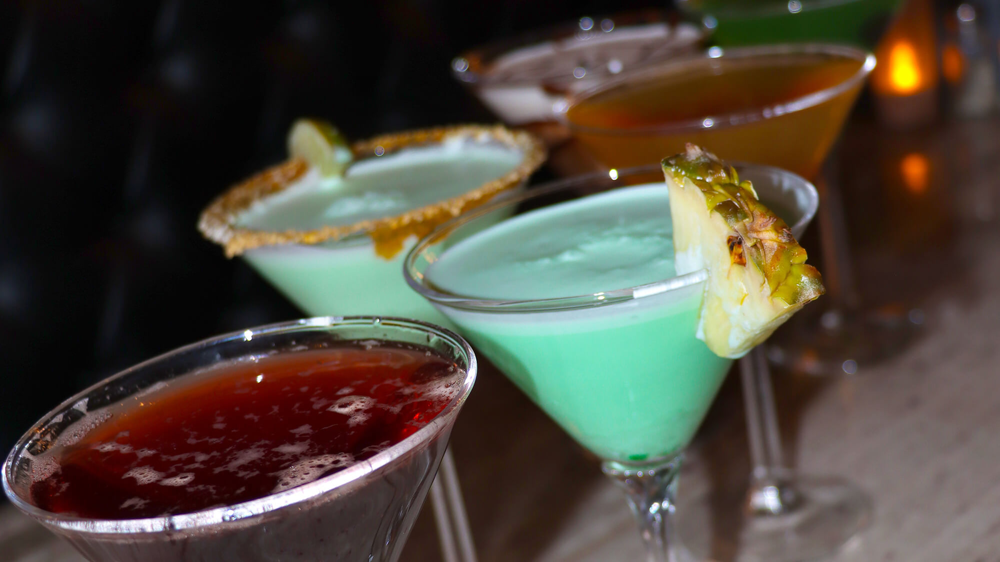 A close-up, angled lineup of multiple colorful martinis on a bar, featuring a dark red cocktail in the front, followed by a bright green martini with a pineapple wedge, and a light green martini with a crumb rim.