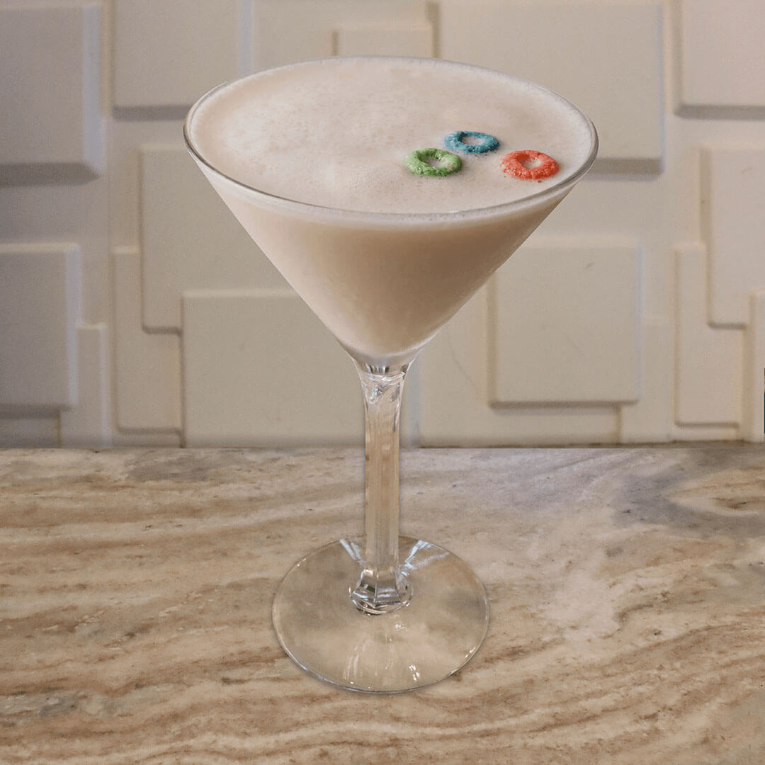 A creamy, pale pinkish-white cocktail garnished with a colorful floating ring of fruit cereal pieces.