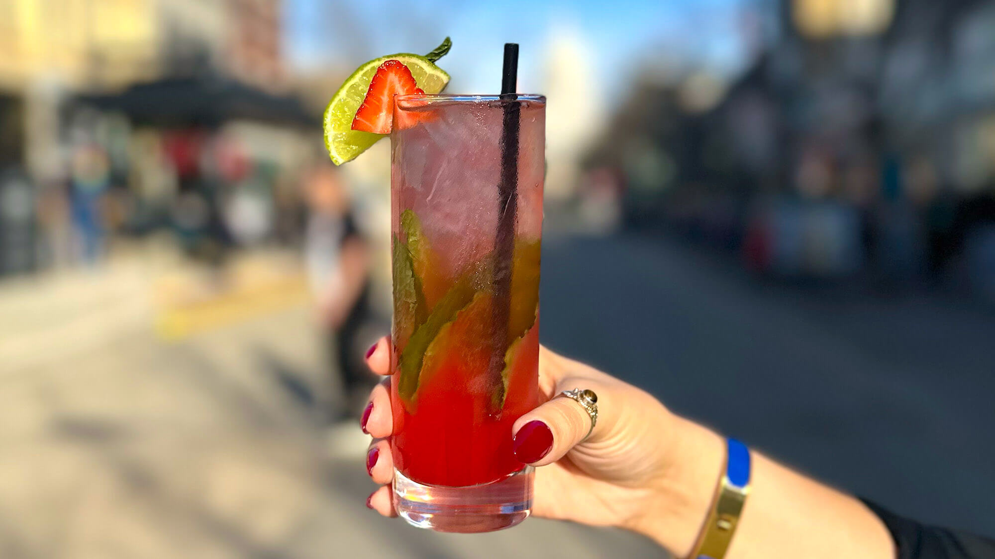 A hand holding a tall, refreshing red cocktail filled with ice and fresh mint leaves, garnished with a lime wedge and a strawberry slice against a blurred outdoor background.