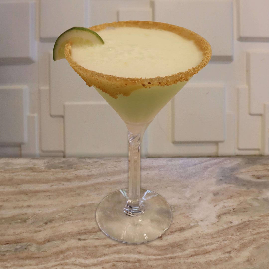 A creamy, pale green lime cocktail featuring a caramel graham cracker rim and a dollop of white whipped cream.