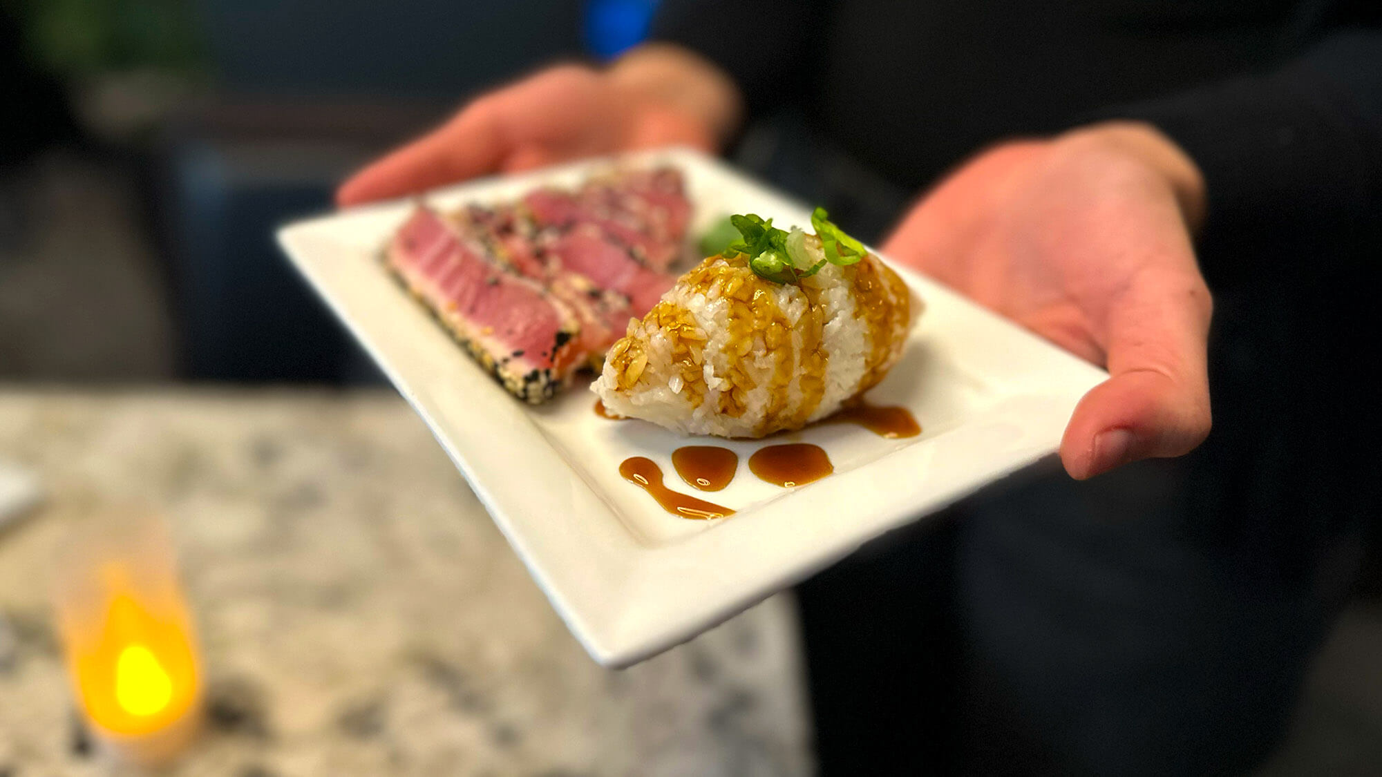 Server holding a small white square plate featuring slices of sesame-crusted seared tuna alongside a portion of rice drizzled with dark savory sauce and topped with green scallions.