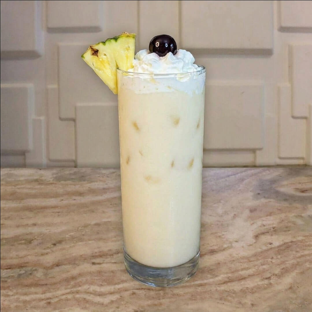 A tall, creamy white frozen mocktail served in a highball glass, topped with whipped cream, a fresh pineapple wedge, and a dark cherry.