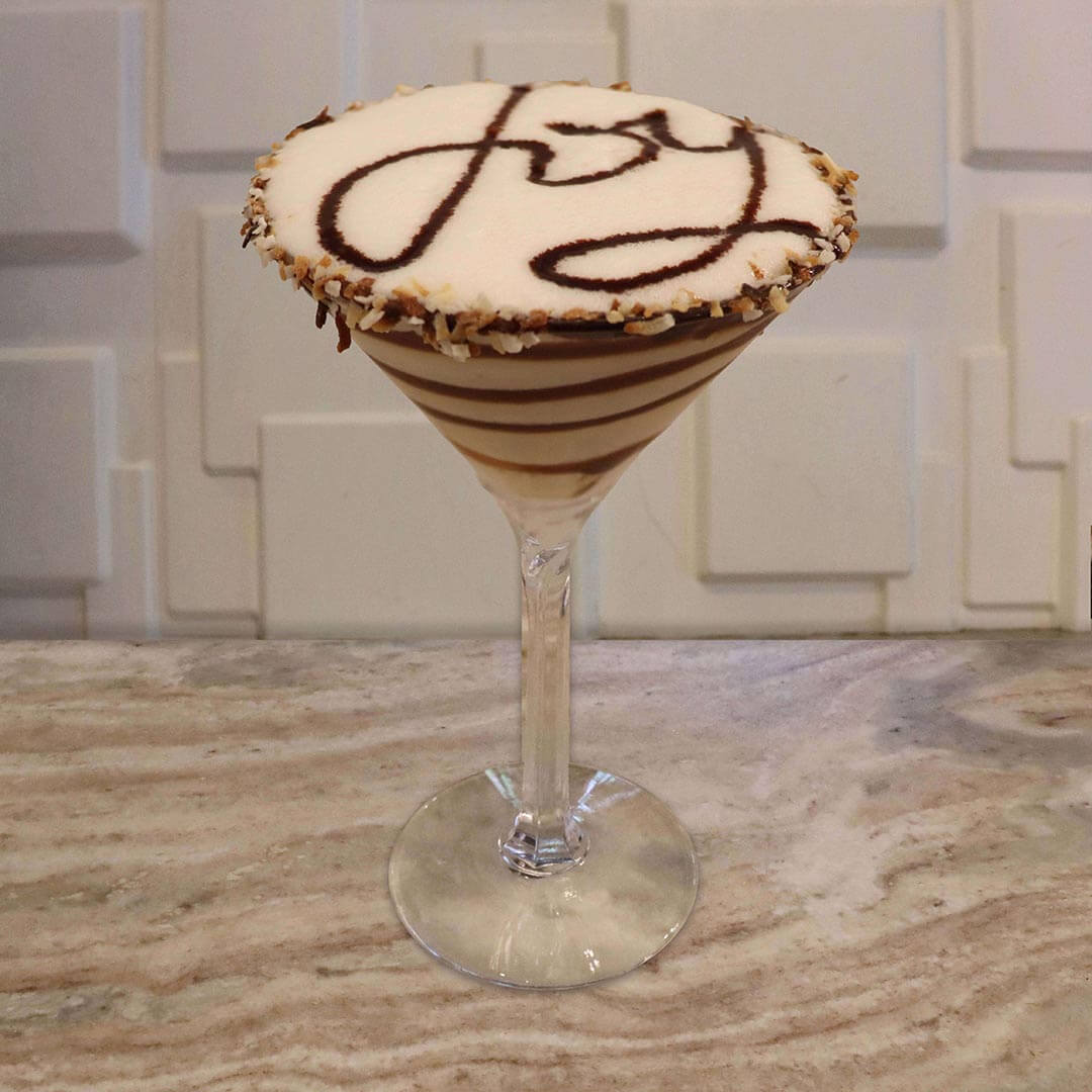 A creamy, white cocktail with thick chocolate syrup swirls inside the glass and a toasted coconut-coated rim.