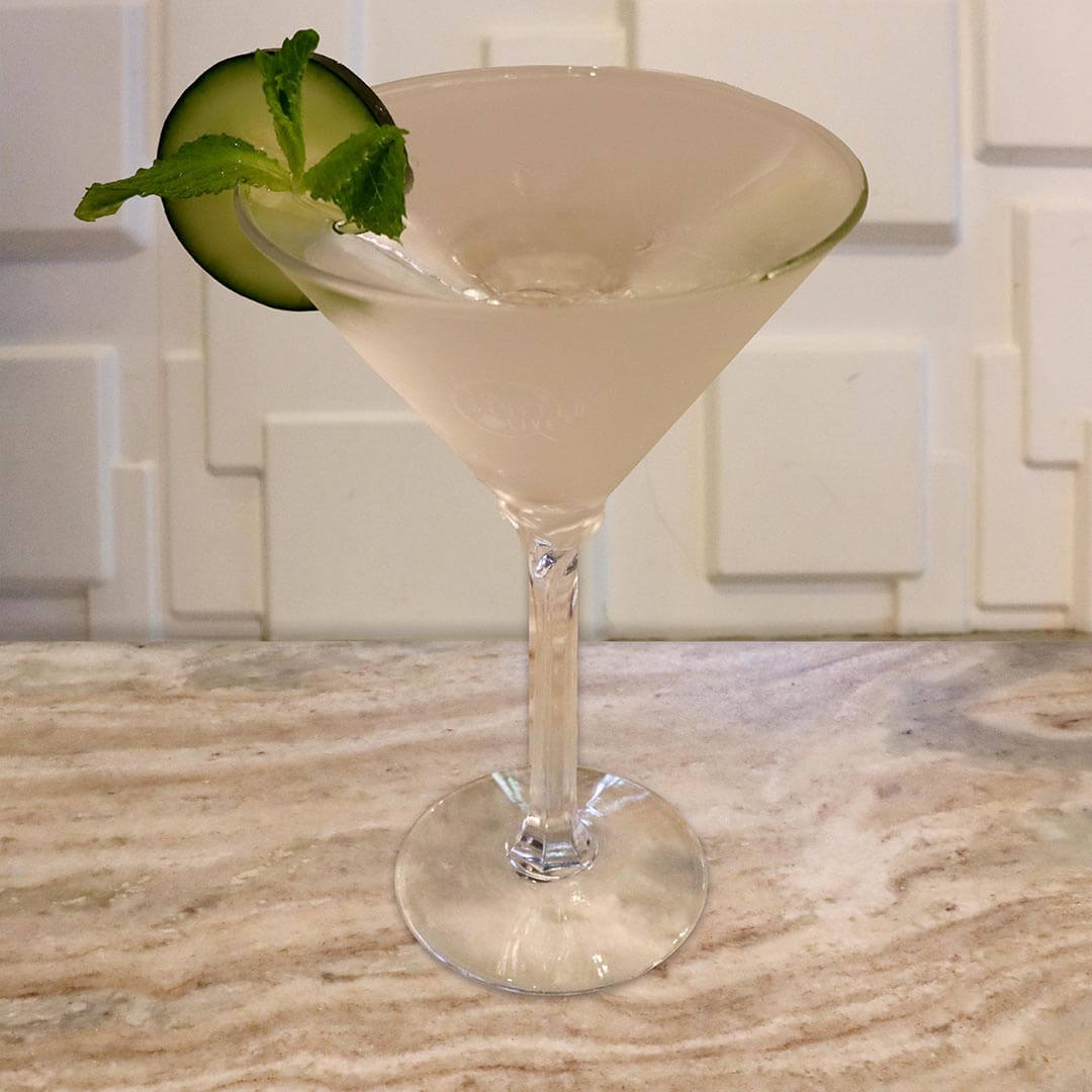 A clear, effervescent cocktail with muddled green mint leaves, garnished with a cucumber slice.