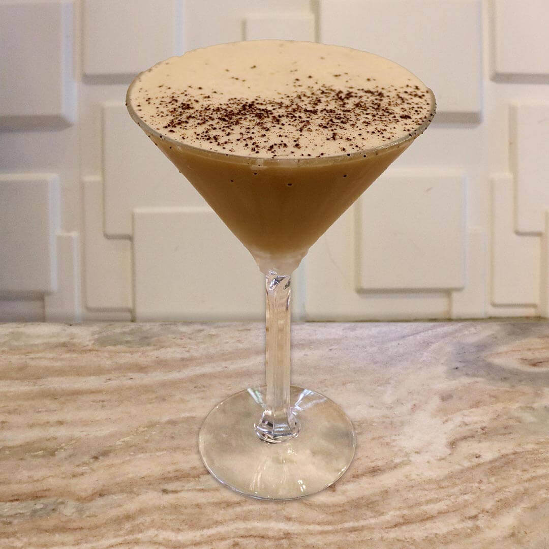 A creamy, light brown espresso cocktail topped with coffee bean grounds, featuring a caramel-drizzled glass.