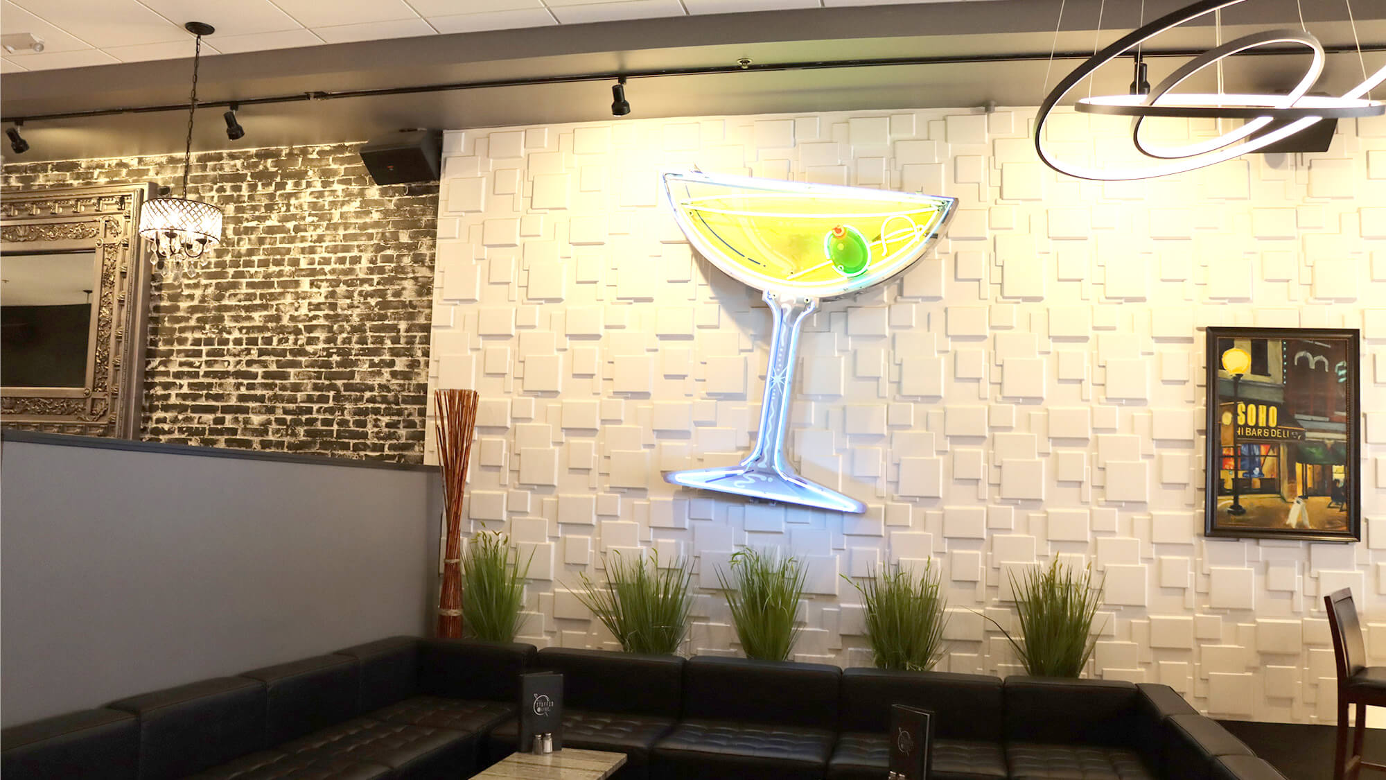 Close-up view of the iconic large glowing neon martini glass sign with a green olive, mounted on a modern geometric patterned wall in the lounge of The Stuffed Olive, Cedar Falls, Iowa.