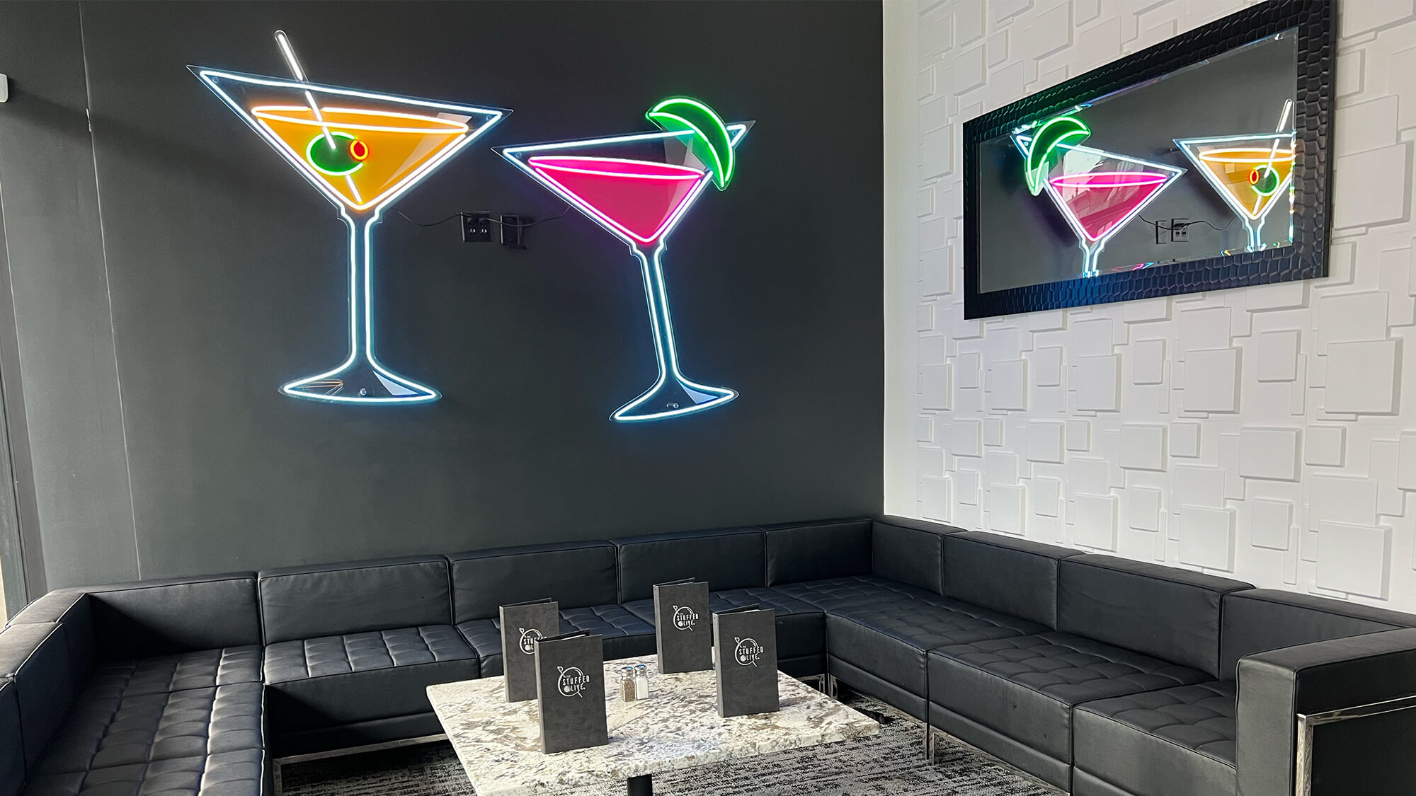 Cozy lounge area at The Stuffed Olive in Omaha featuring a large black leather sectional sofa, a marble table with menus, a textured white accent wall, and bright neon martini signs.