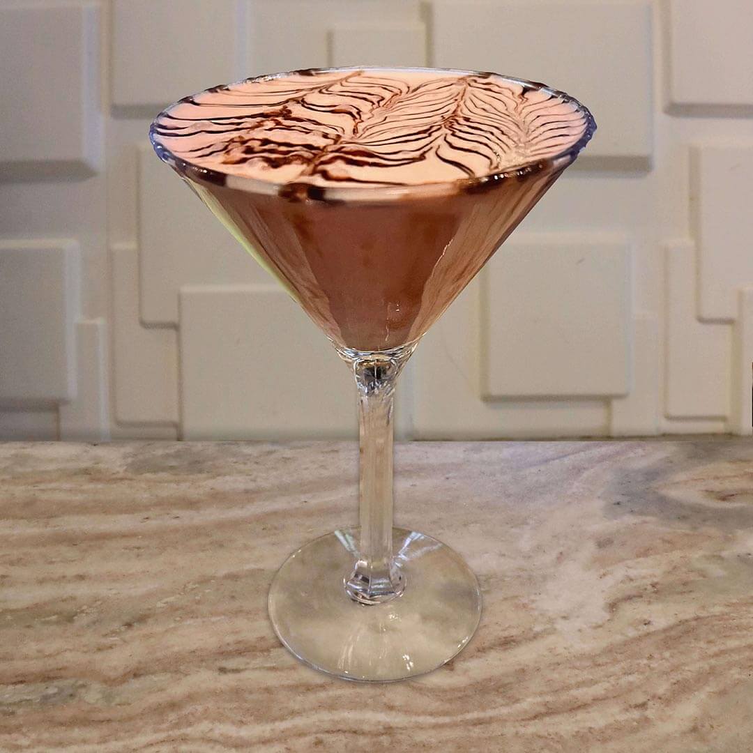 A creamy pink cocktail in a glass with a chocolate-coated rim, featuring an intricate, feathered chocolate syrup design across the top.