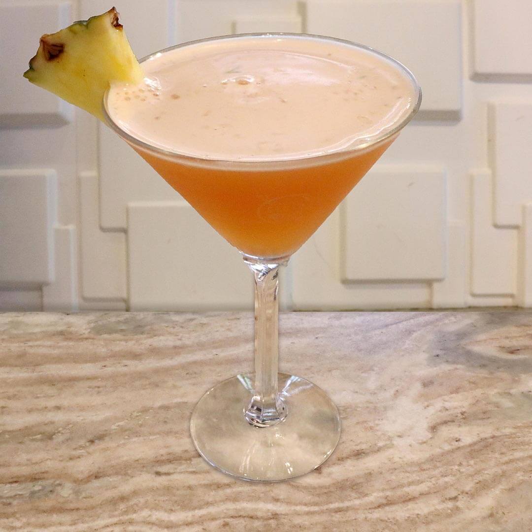 A hazy, golden-orange tropical cocktail served in a chilled glass, garnished with a fresh pineapple wedge.