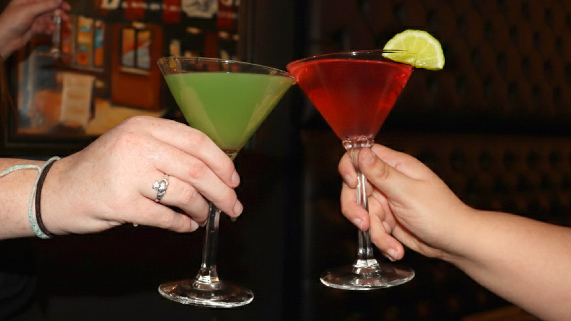 Close-up of two hands clinking a vibrant green martini and a bright red martini garnished with a lime wedge in a dimly lit lounge setting.