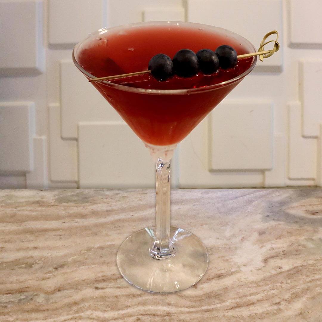 A deep, vibrant red cocktail in a chilled glass, garnished with fresh blueberries on a pick.