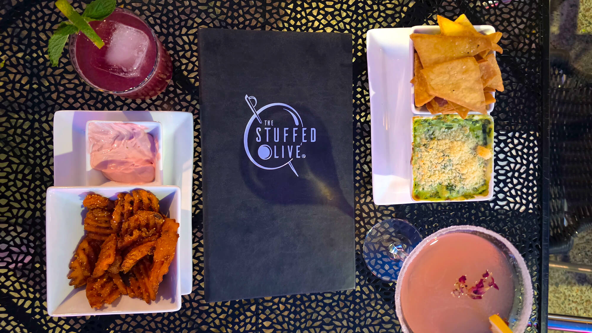 Overhead view of a black patio table featuring The Stuffed Olive menu, surrounded by two colorful craft cocktails, sweet potato waffle fries, and two square plates of creamy dips with pita points.
