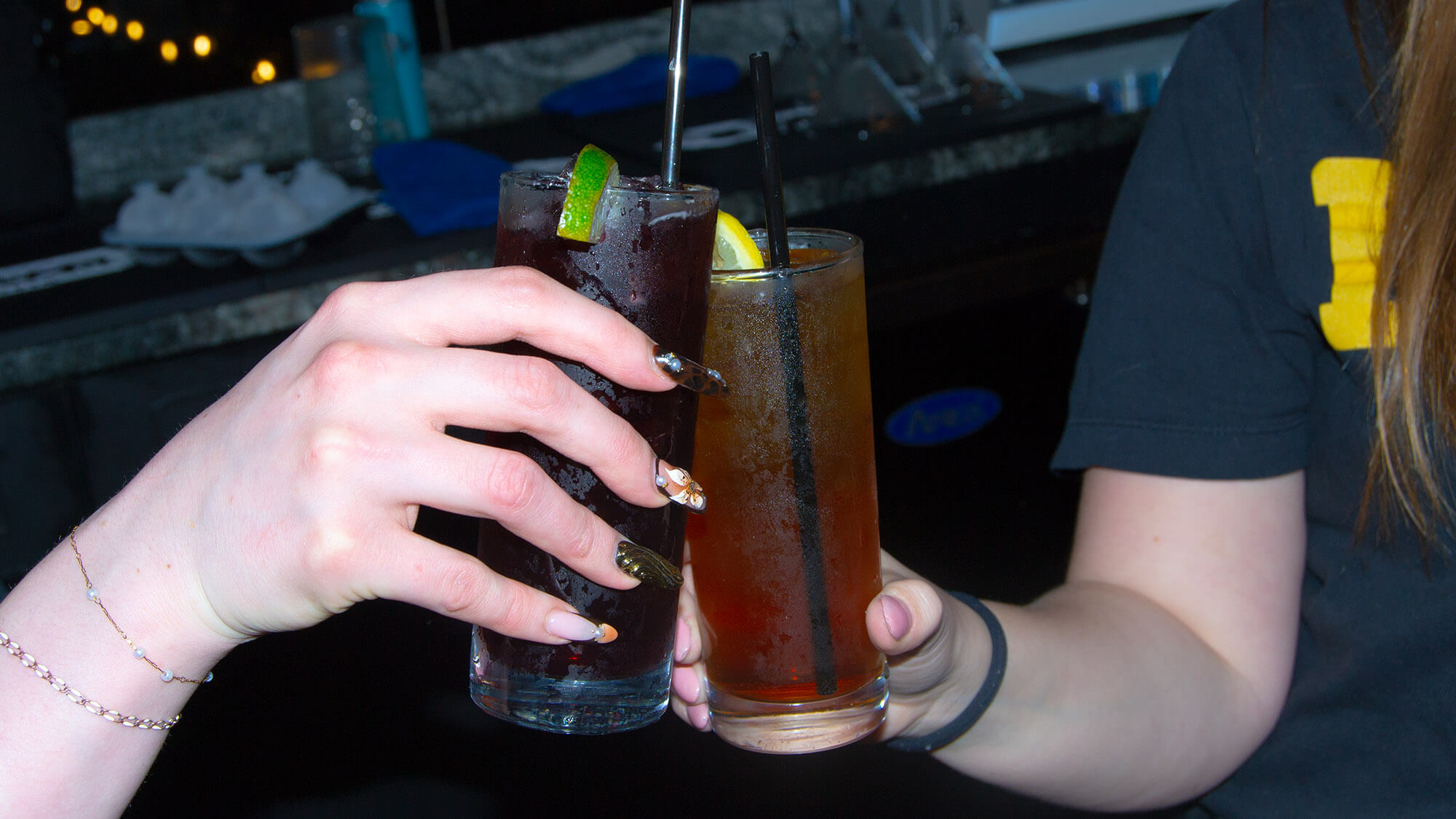Two people clinking tall mixed drinks together at a bar. One glass holds a dark iced cocktail garnished with a lime, and the other holds an amber iced drink garnished with a lemon.