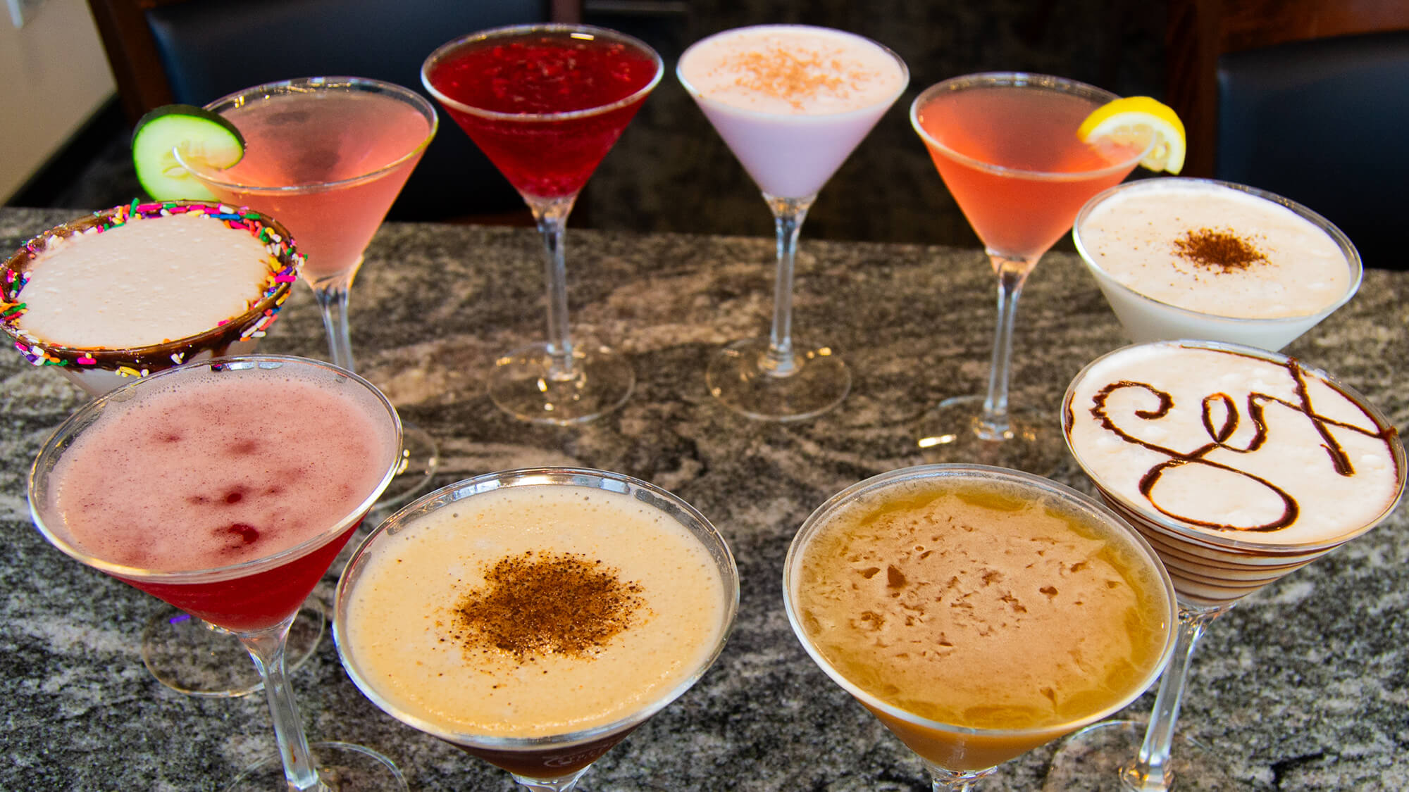 Nine colorful signature martinis arranged in a circle on a granite bar top, featuring a variety of flavors, colors, and garnishes including sprinkles, chocolate drizzle, and fresh fruit.