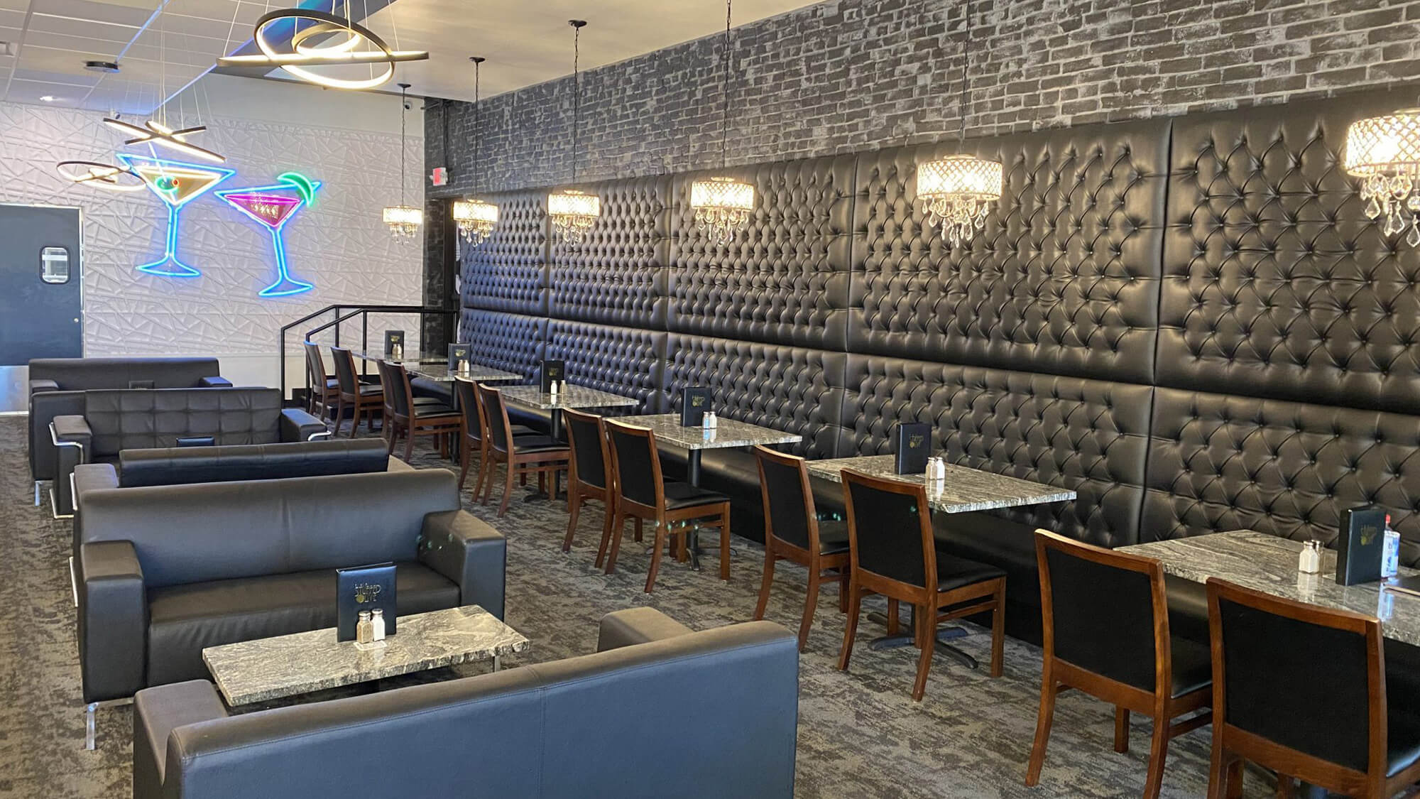 Modern lounge area at The Stuffed Olive in Iowa City, featuring black tufted leather seating, marble tables, and a vibrant neon sign with two martini glasses.