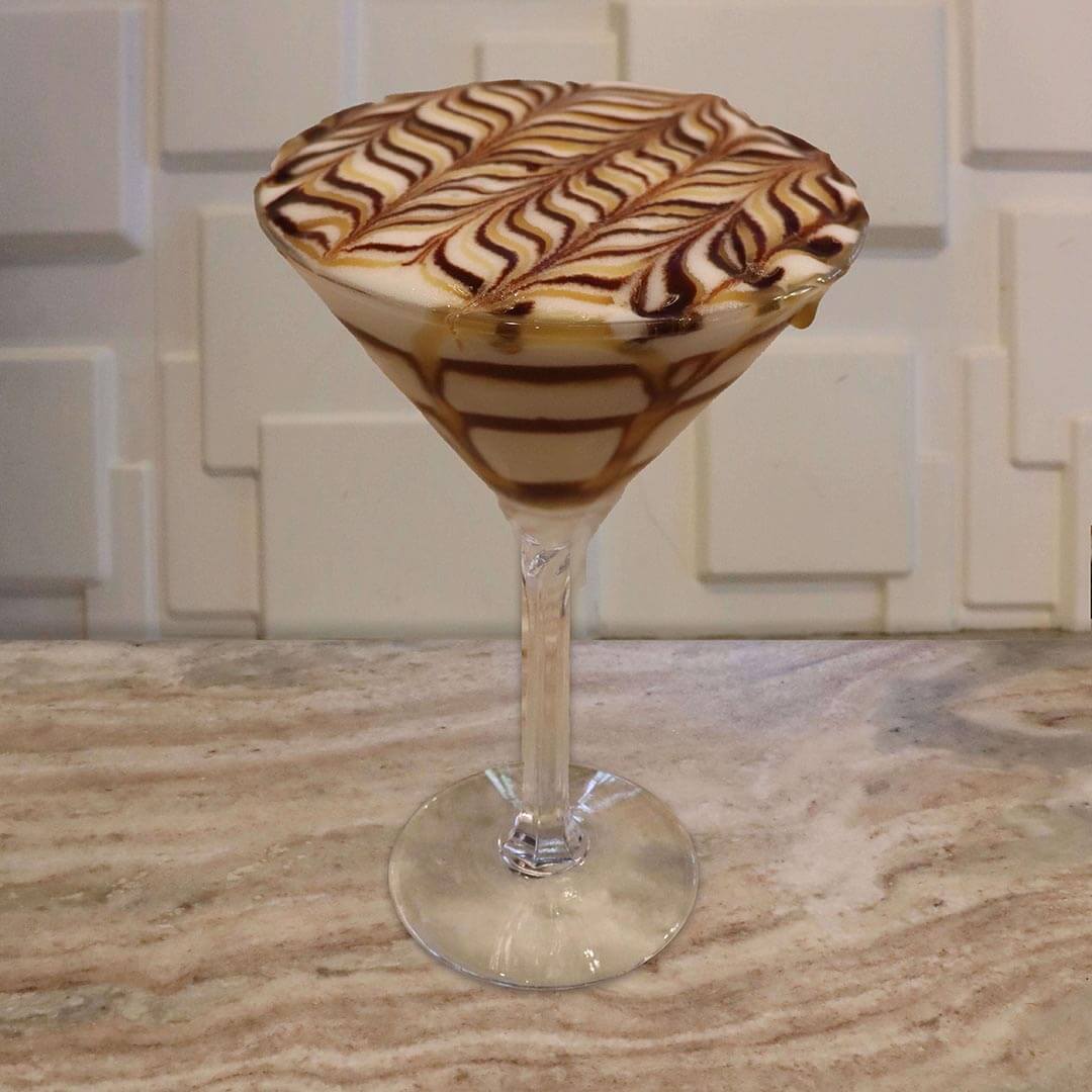 A creamy, light brown cocktail featuring intricate chocolate and caramel swirls coating the inside of the glass.