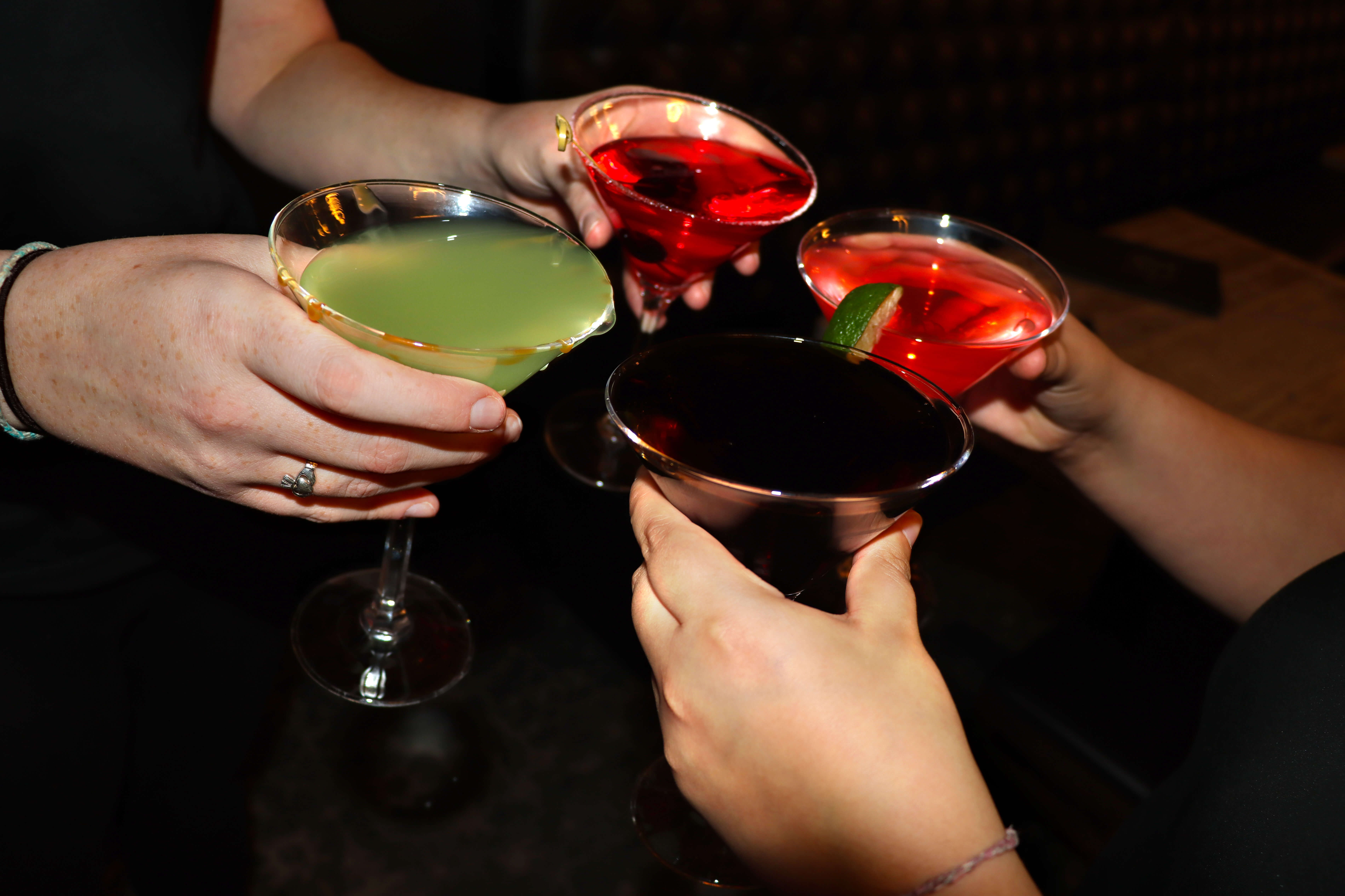A close-up photograph of four people holding and clinking different colored martini glasses in a toast. The cocktails are green, dark red, and two vibrant reds, held against a dimly lit bar background.